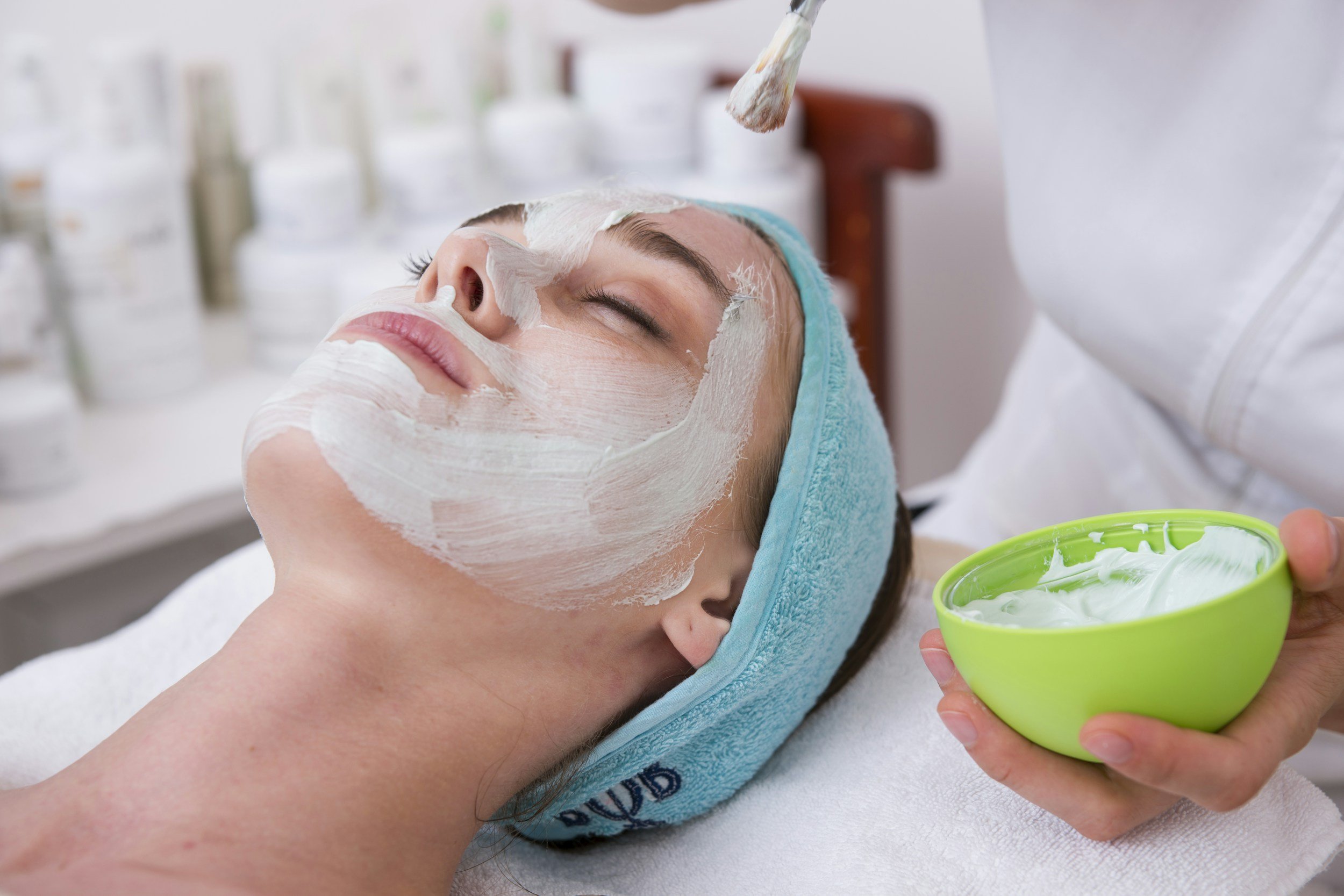 Person receiving facial treatment with cream applied on face, wearing a blue headband, in a skincare setting.
