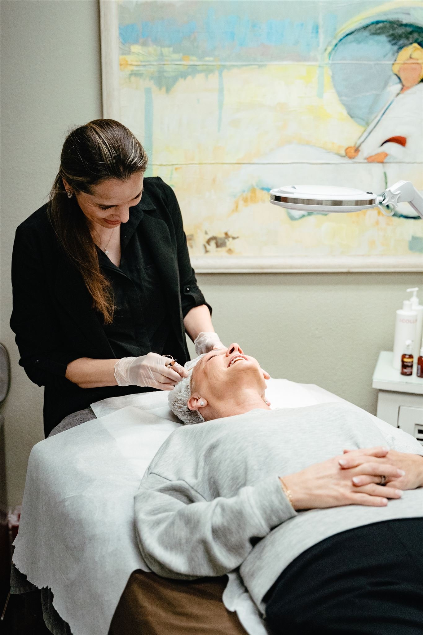 A woman undergoing a facial or cosmetic treatment in a spa or clinic, lying on a bed while another woman, wearing gloves and using a special light, performs the procedure. The setting is professional with spa or clinical equipment visible.