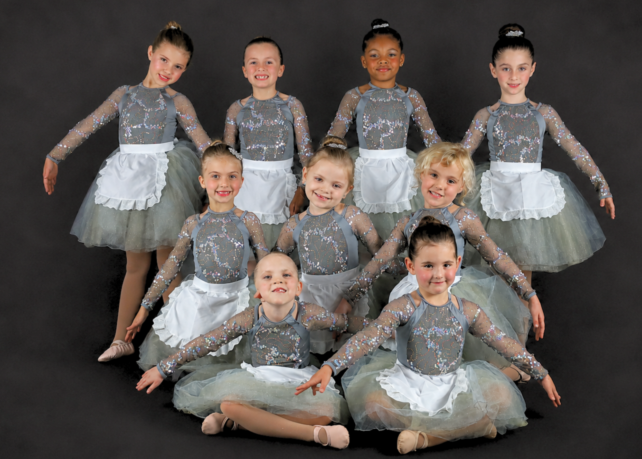 Group of nine young girls dressed in purple ballet tutus and leotards, posing together on a black floor against a white background, each with hands crossed over their chests.