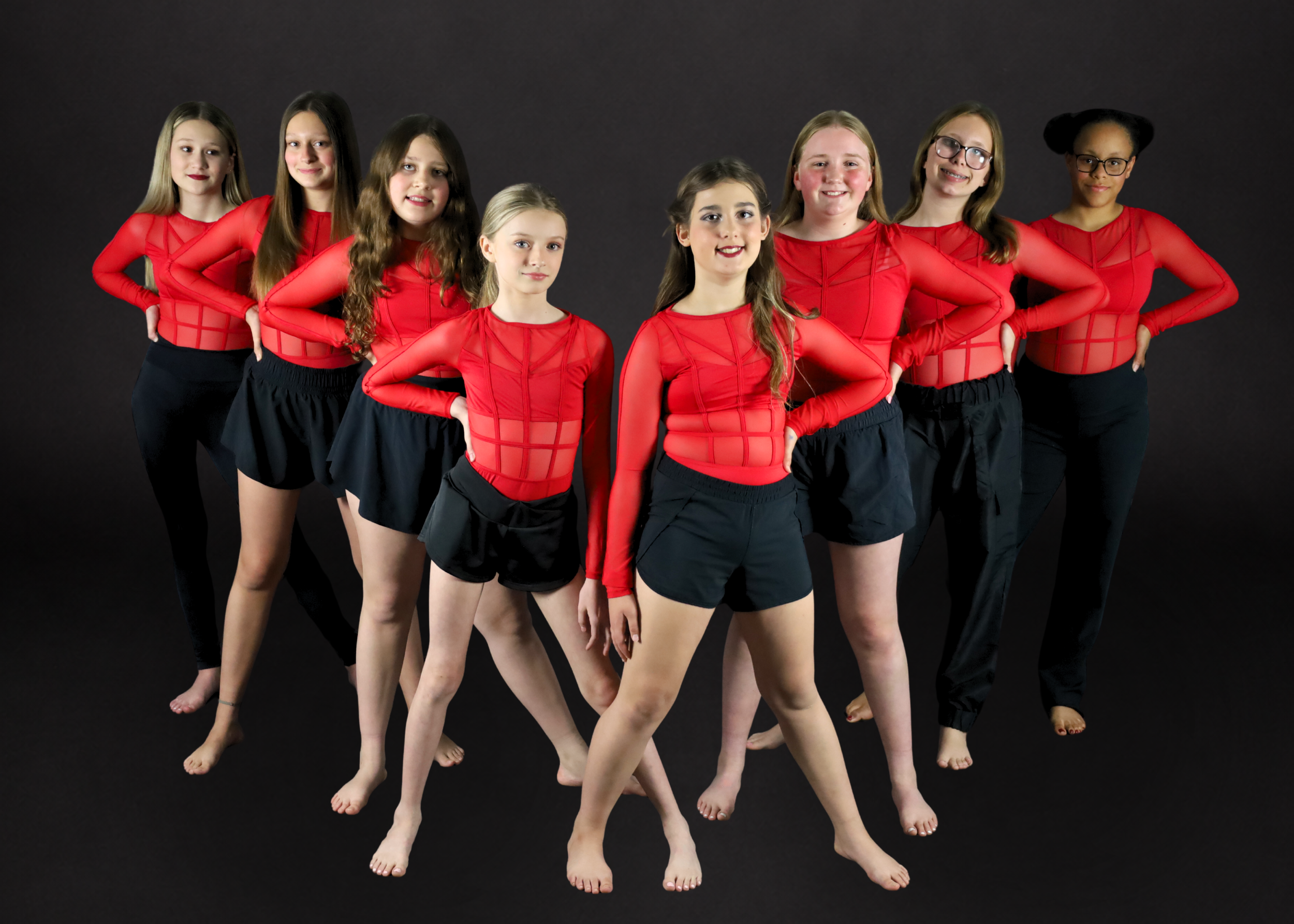 Group of ten young girls wearing matching burgundy dance dresses, posing on black floor with a white background.