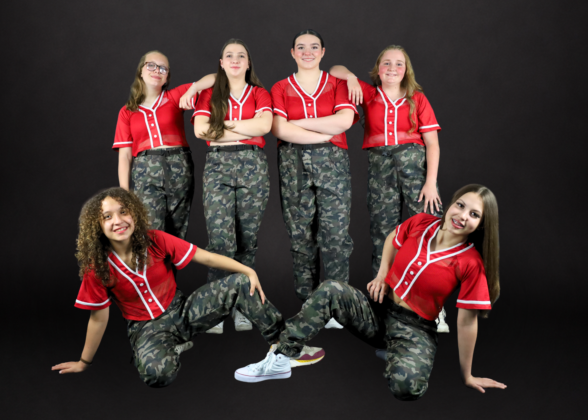 Eight young women in matching gray and black dance costumes pose for a group photo against a white background, with some sitting, kneeling, and standing.