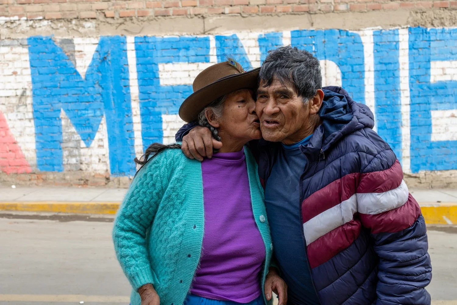 A woman in a brown hat kisses a man’s cheek as they embrace outside a brick wall in Lima