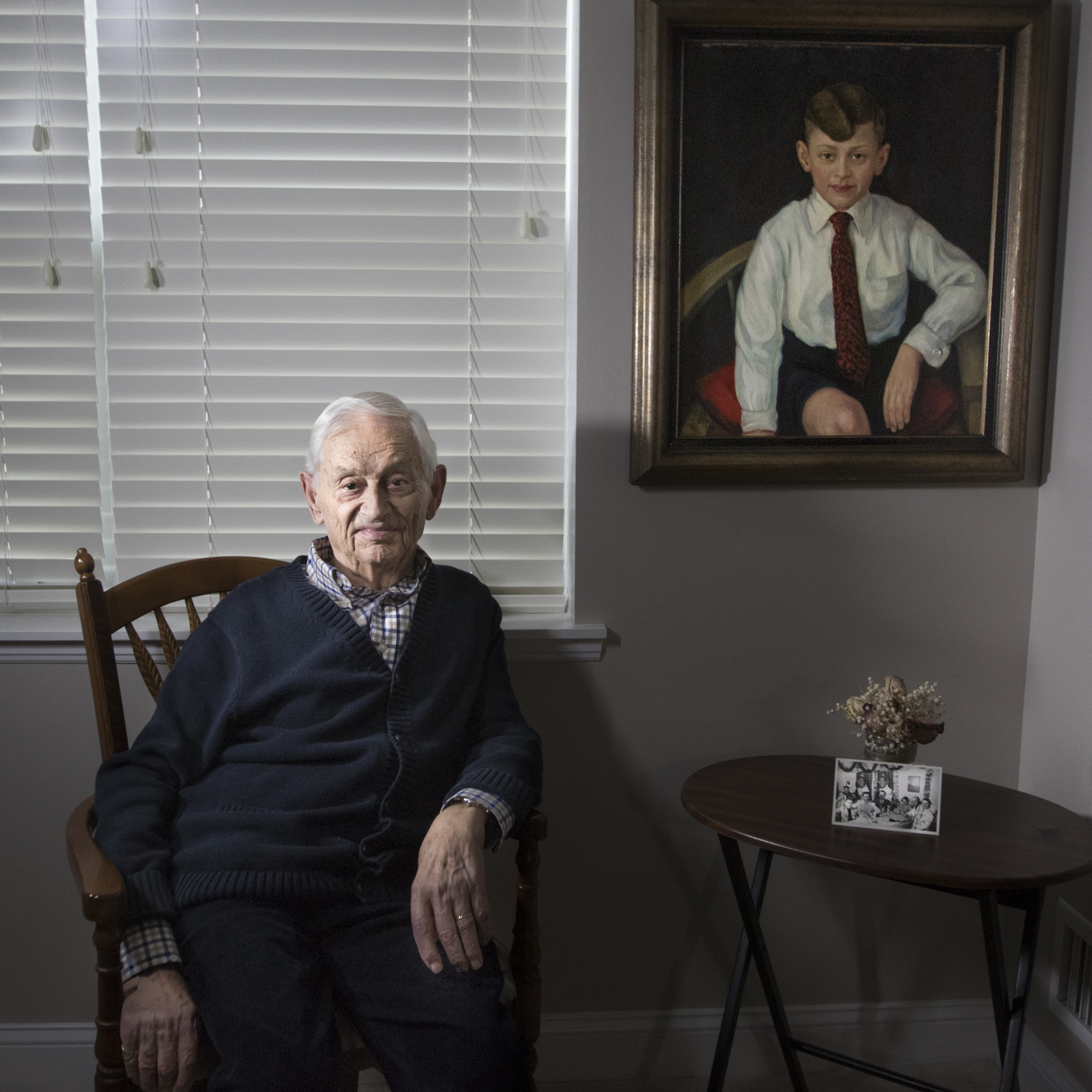Egon Salmon poses next to a painting of him which was made in Rheydt, Germany in 1933, when Salmon was 9 years old. The portrait traveled with him on the S.S. St. Louis, to Belgium, and eventually to the United States in 1940. The small photo leaning against the flowers pictures him with his mother, sister, and other passangers at his table on the St. Louis ship. Shortly after his arrival to the United States, he settled with his family on Staten Island and was drafted to fight overseas in the American Army.