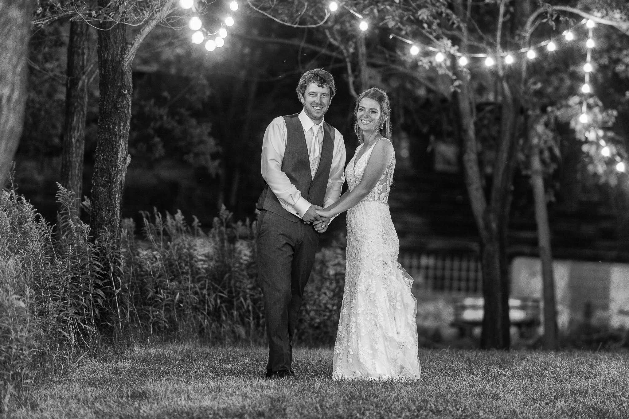 Black and white night portrait under string lights draped through trees, groom in vest and dress shirt, bride in lace gown, dark garden setting — Milwaukee wedding photography