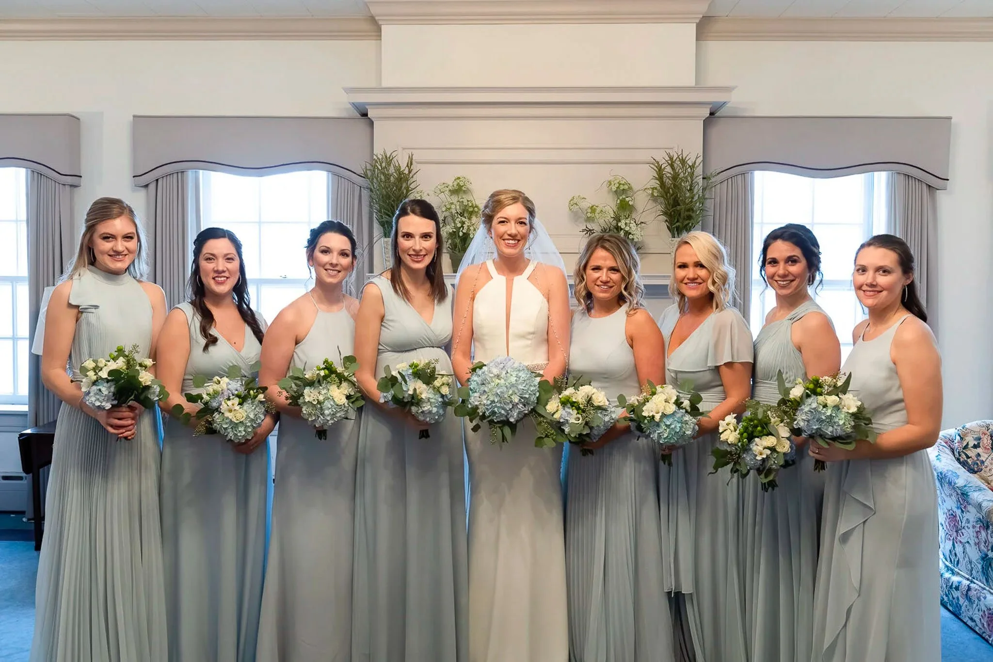 Bride surrounded by eight bridesmaids in sage-green gowns holding blue hydrangea and white rose bouquets, elegant formal venue interior with large windows — Milwaukee wedding photography