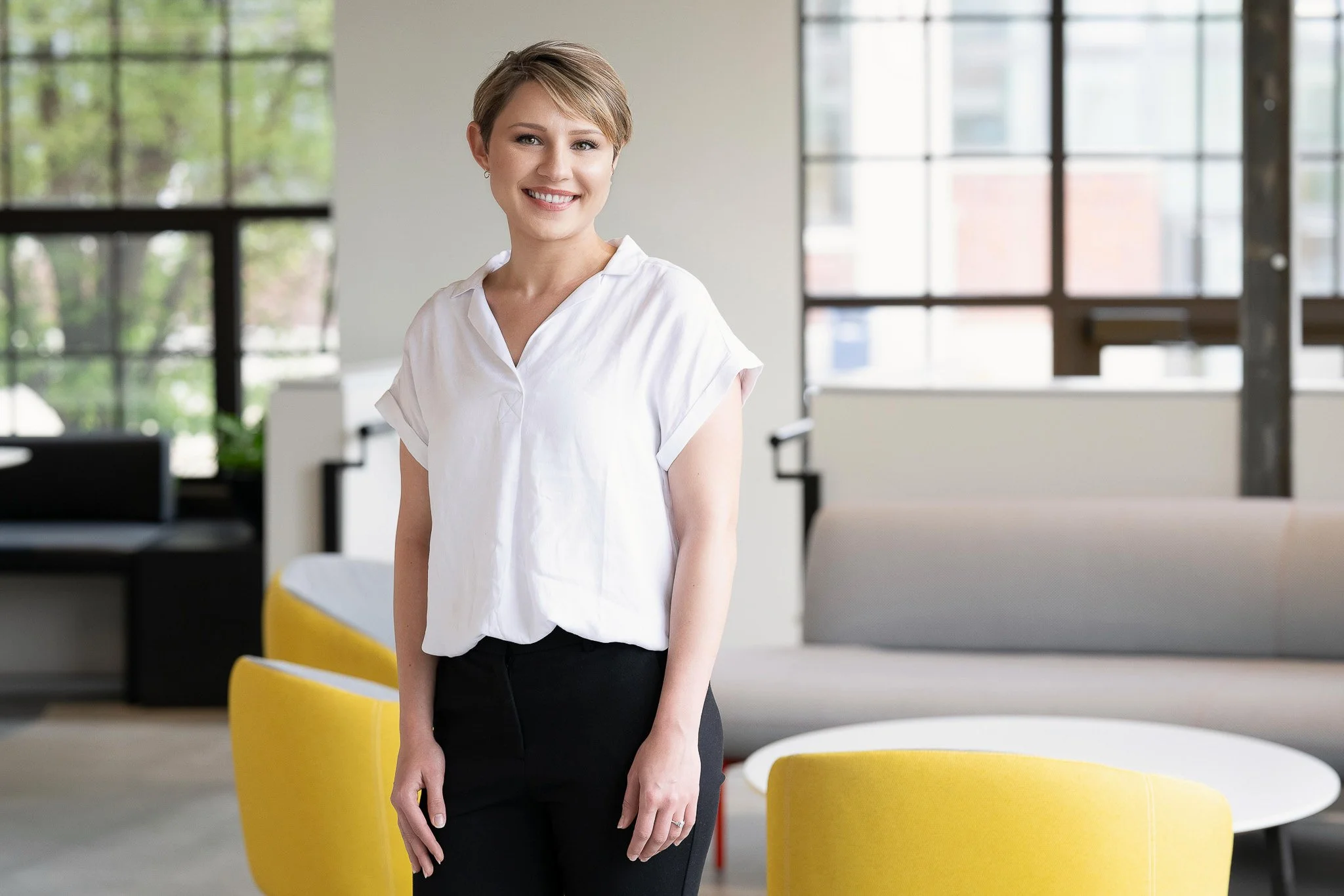Professional woman headshot, short blonde hair, white short-sleeve top, standing in modern corporate lobby with yellow accent chairs and large industrial windows behind — Milwaukee corporate headshot photography