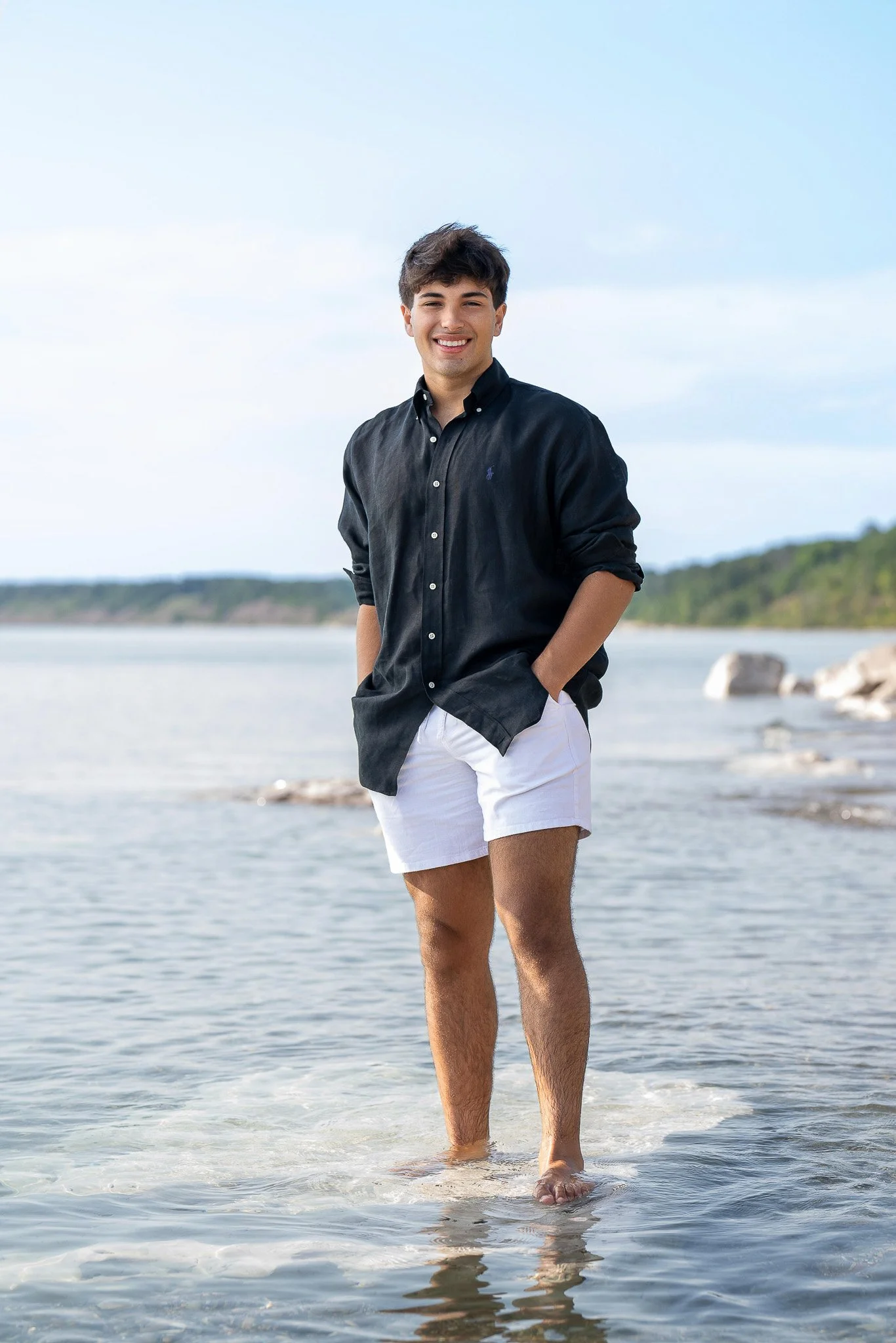 Teen boy standing barefoot in shallow lake water, black shirt and white shorts, smiling broadly with rocky shoreline and distant treeline behind — Milwaukee senior portrait photography