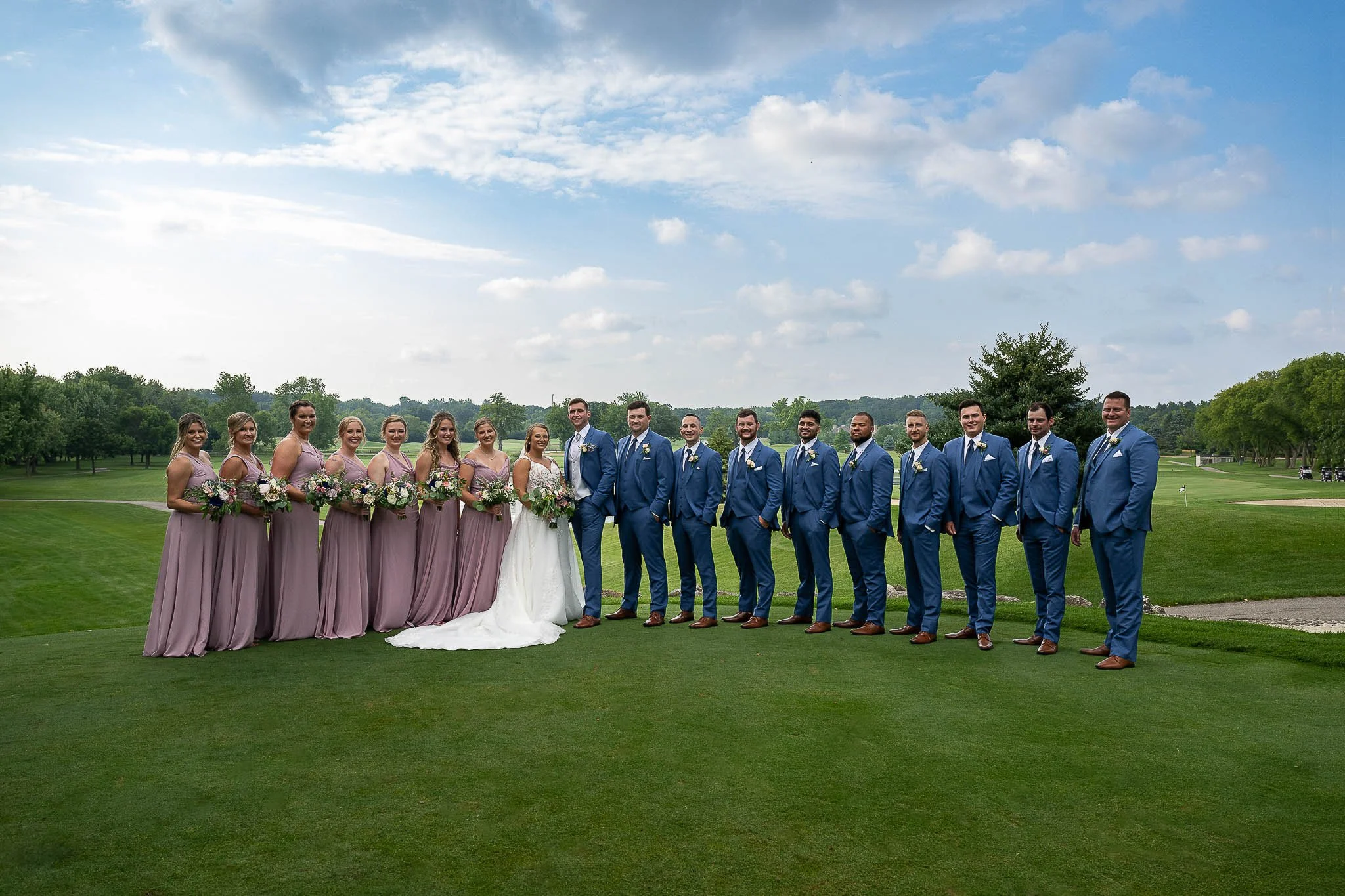 Full wedding party lined up on golf course fairway, mauve dusty rose bridesmaids with mixed bouquets, groomsmen in navy blue suits, dramatic cloudy sky — Milwaukee wedding photography