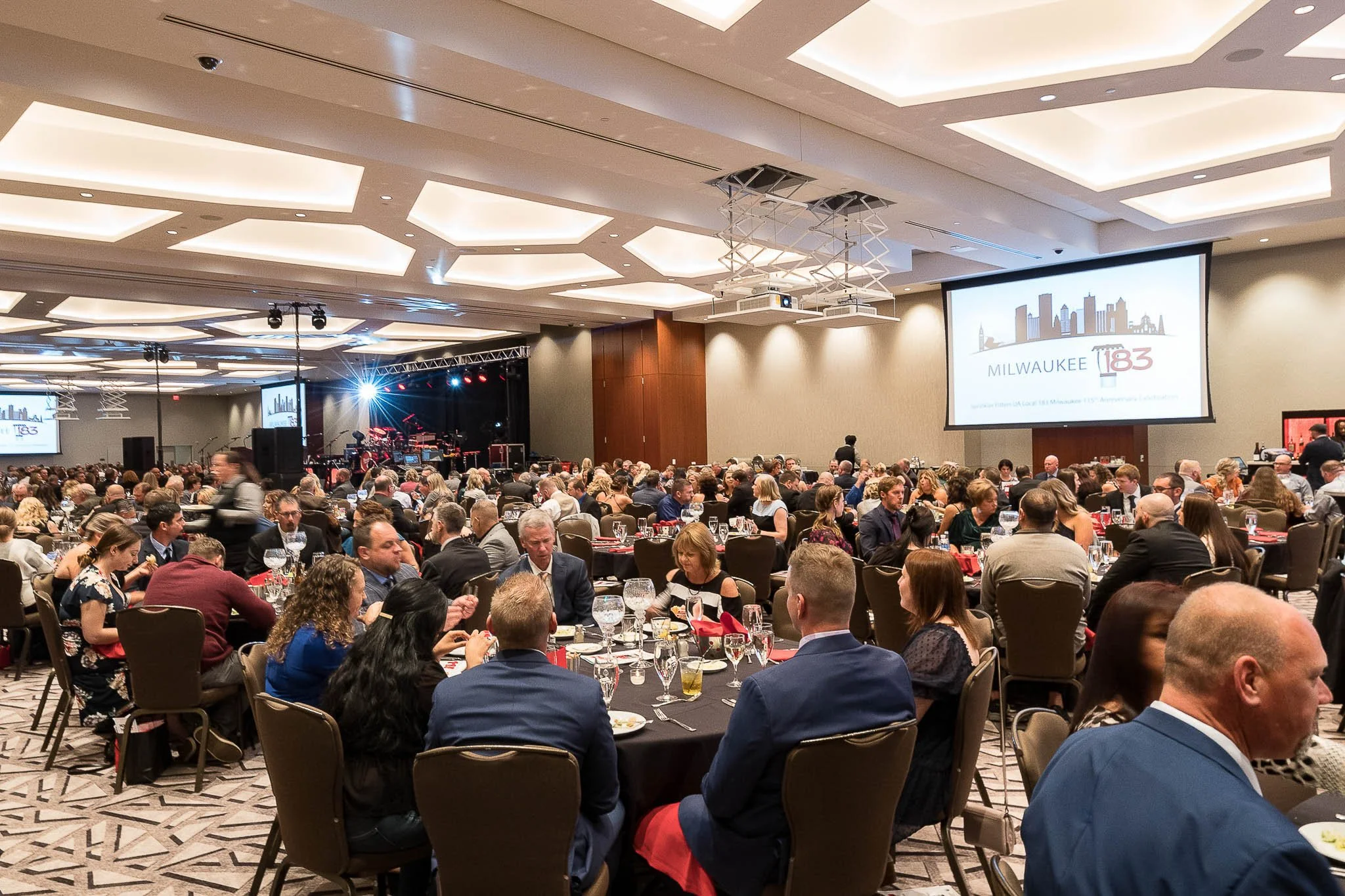 Large hotel ballroom gala with hundreds of guests at black-linened round tables, "Milwaukee 1183" on projection screens, stage with band setup in background — Milwaukee Sprinkler Fitters Local 183 event photography