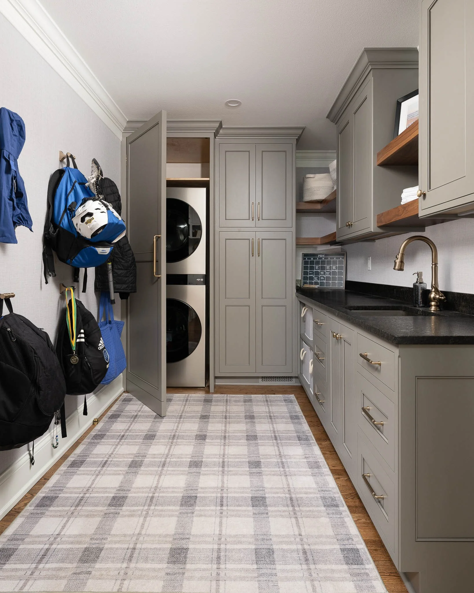 Mudroom and laundry room with grey-green shaker cabinets, stacked washer and dryer, plaid rug, brass hardware, and backpack hooks — Milwaukee residential interior photography
