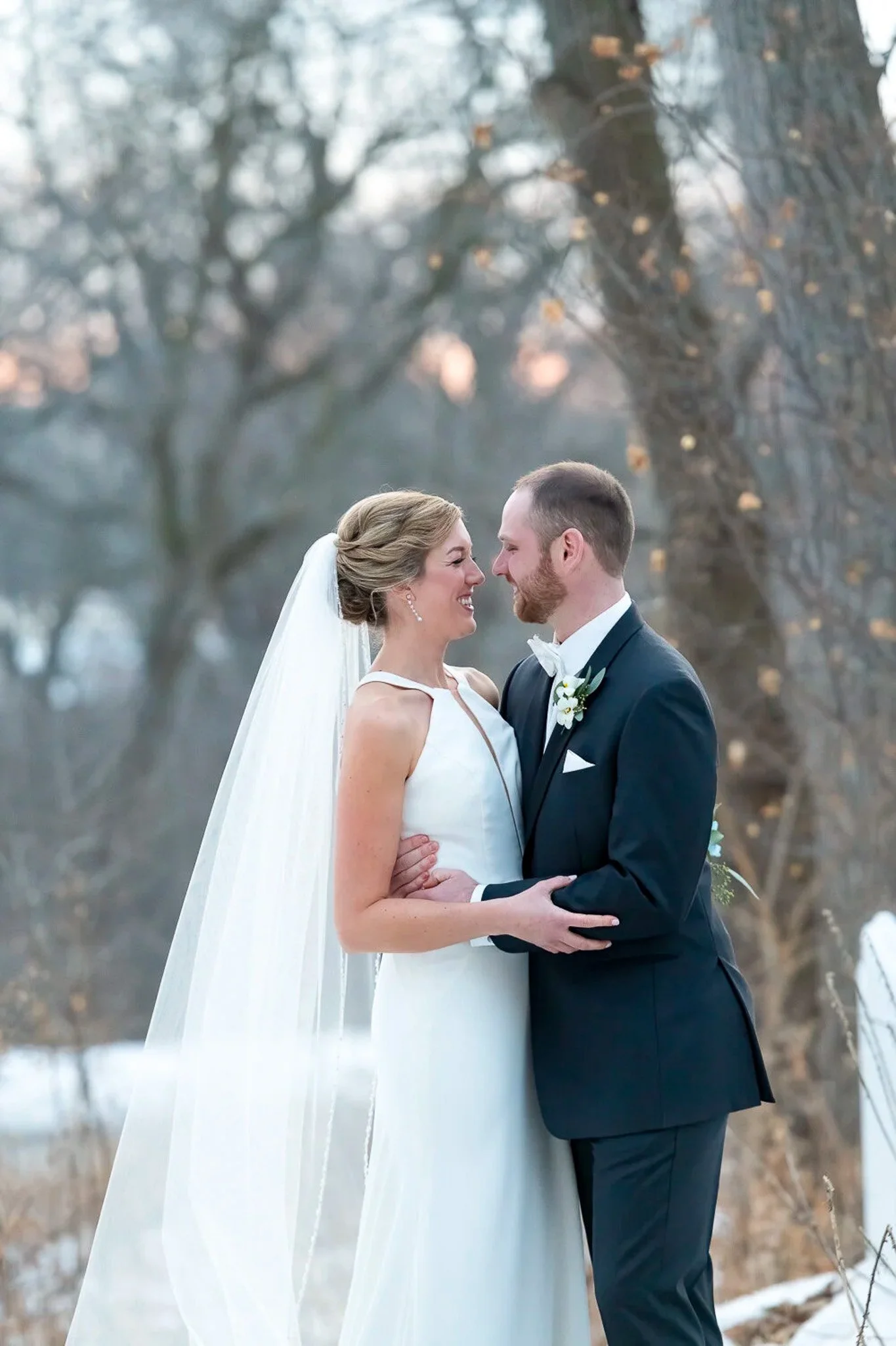 Winter couple portrait in snow, bride in simple white halter gown with cathedral veil, groom in dark navy suit, bare trees with warm bokeh lights behind — Milwaukee wedding photography