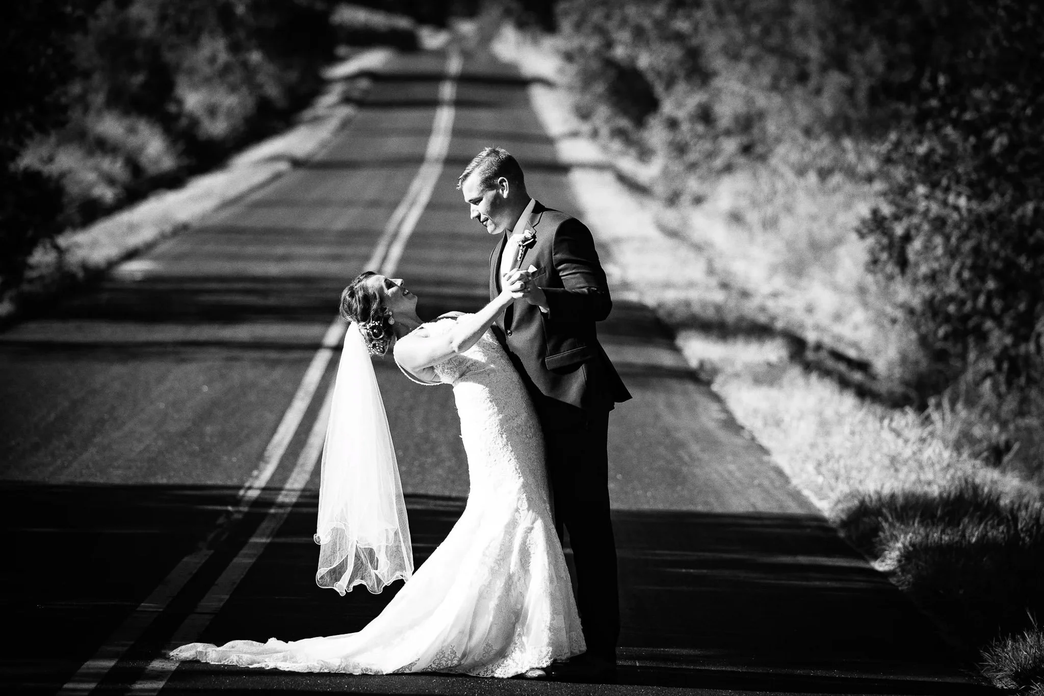 Black and white road dip on winding hilltop country road, groom in dark tuxedo, bride in fitted lace gown with cathedral veil flowing — Milwaukee wedding photography