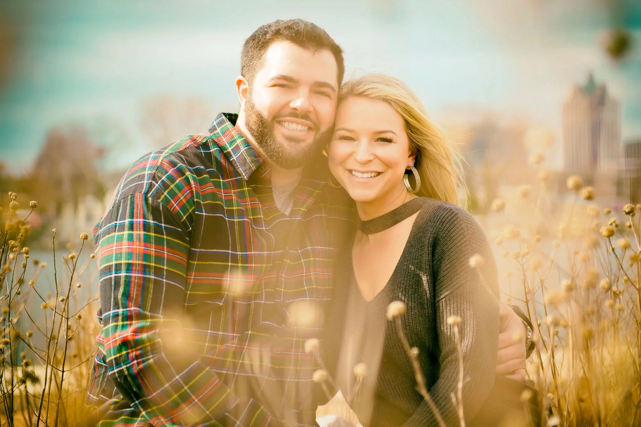 Engagement couple outdoors, man in multicolored plaid flannel, woman in dark grey sweater, Milwaukee skyline softly visible in bokeh background — Milwaukee engagement photography