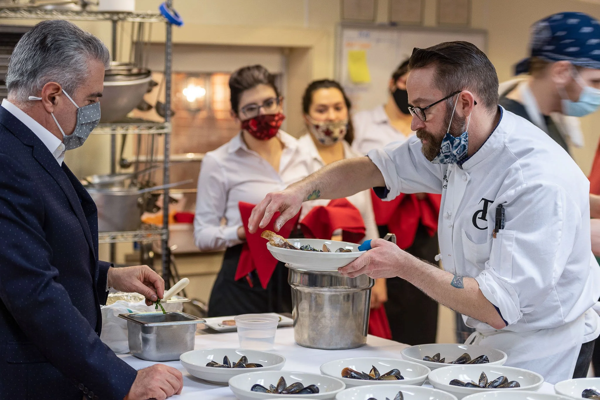 Chefs in white uniforms plating oyster dishes with red bow accents at a catering event, masked guests visible in background — Milwaukee culinary event photography