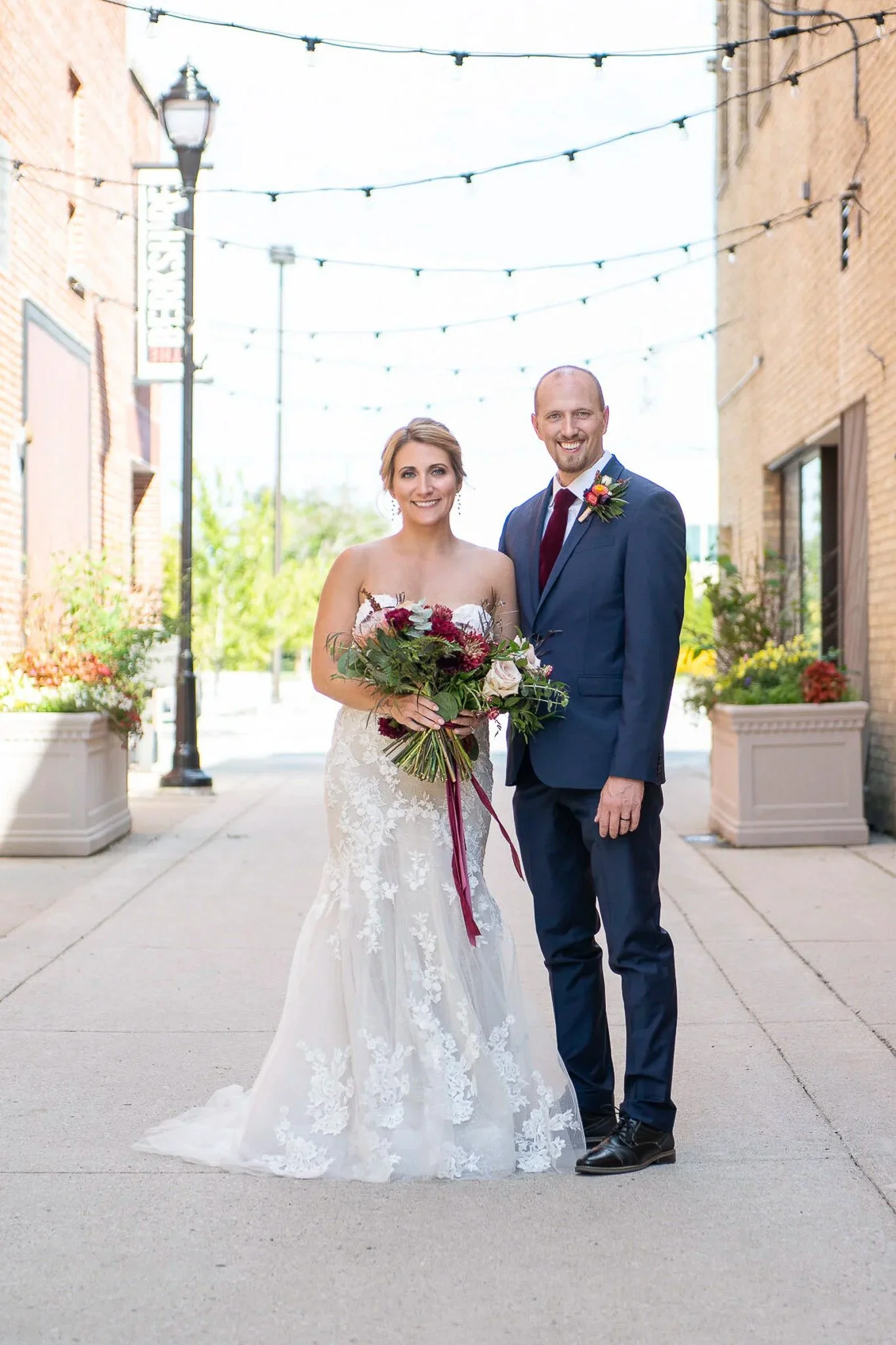 Urban alley couple portrait with string lights overhead, groom in navy suit with burgundy tie, bride holding large burgundy and blush bouquet with ribbon accent — Milwaukee wedding photography