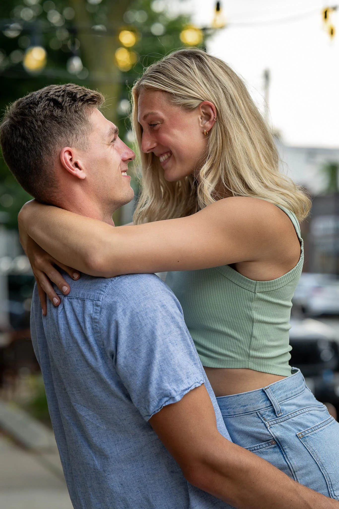 Engagement couple embracing on urban sidewalk, woman lifted wearing sage green ribbed crop top and light jeans, man in light blue shirt, string lights above — Milwaukee engagement photography