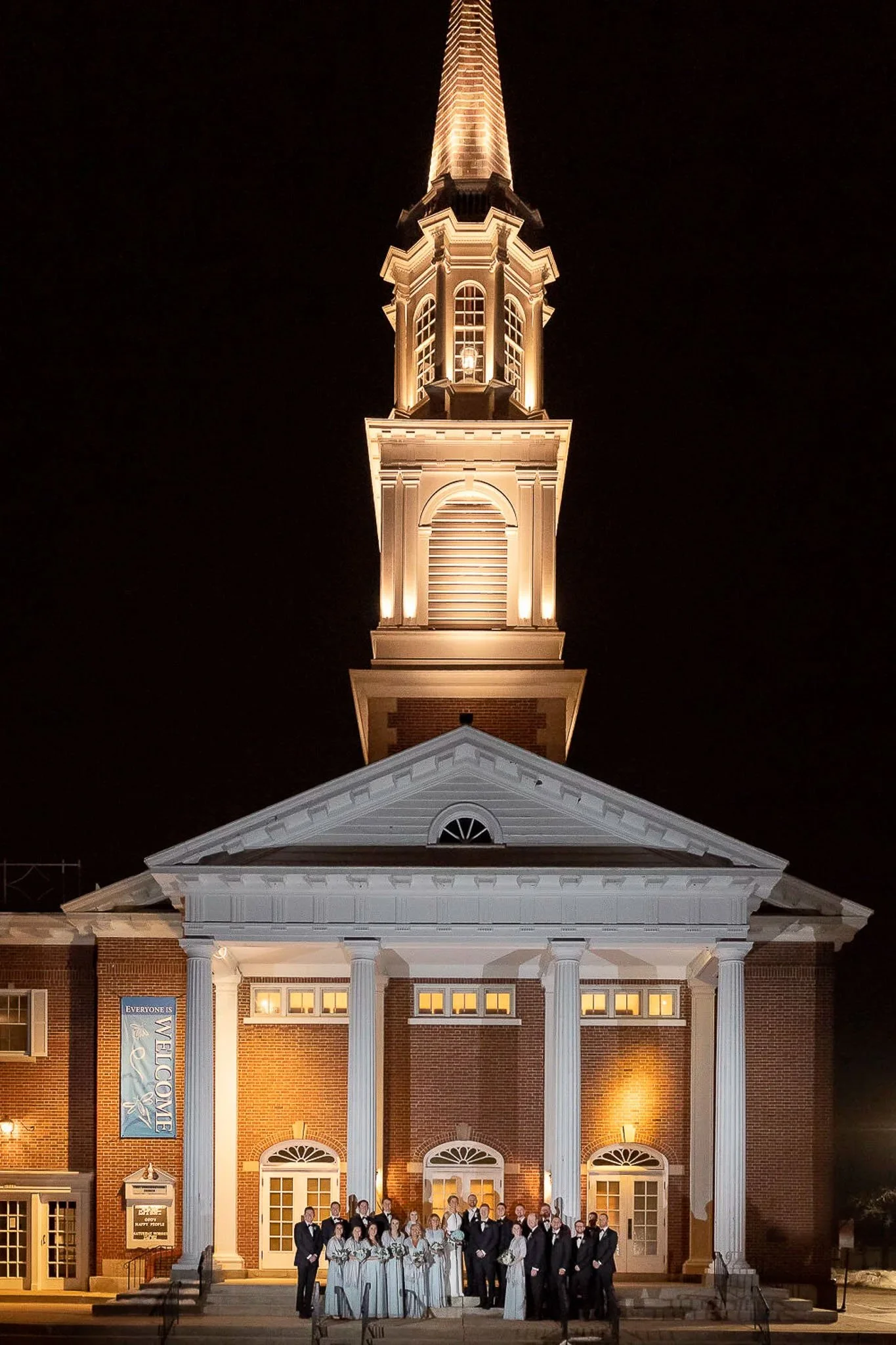 Full wedding party posed in front of illuminated brick church with gold steeple glowing against night sky — Milwaukee wedding photography
