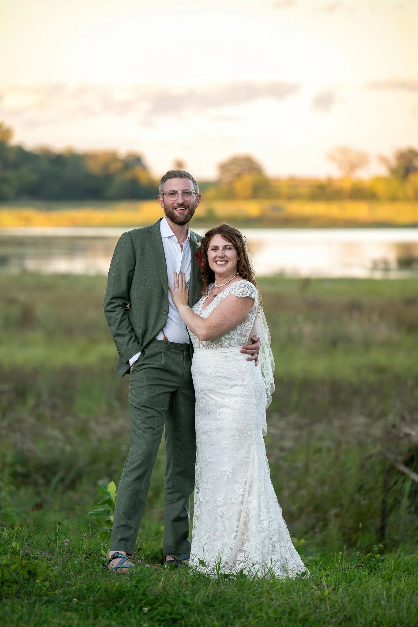 Couple portrait at Wisconsin wetland at sunset, groom in dark green linen suit with glasses, bride in cap-sleeve lace gown with pearl necklace, golden sky reflected in pond — Milwaukee wedding photography