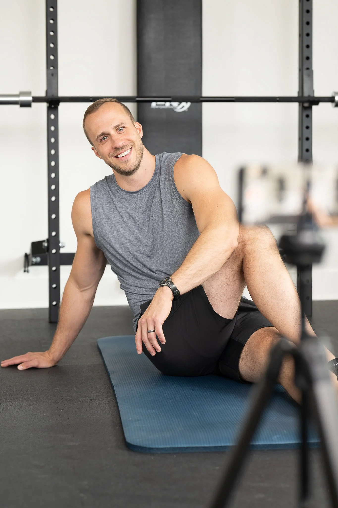 Fitness trainer sitting on blue yoga mat at gym squat rack, grey tank top and black shorts, smiling at camera with squat rack and barbell visible behind — Milwaukee fitness brand portrait photography