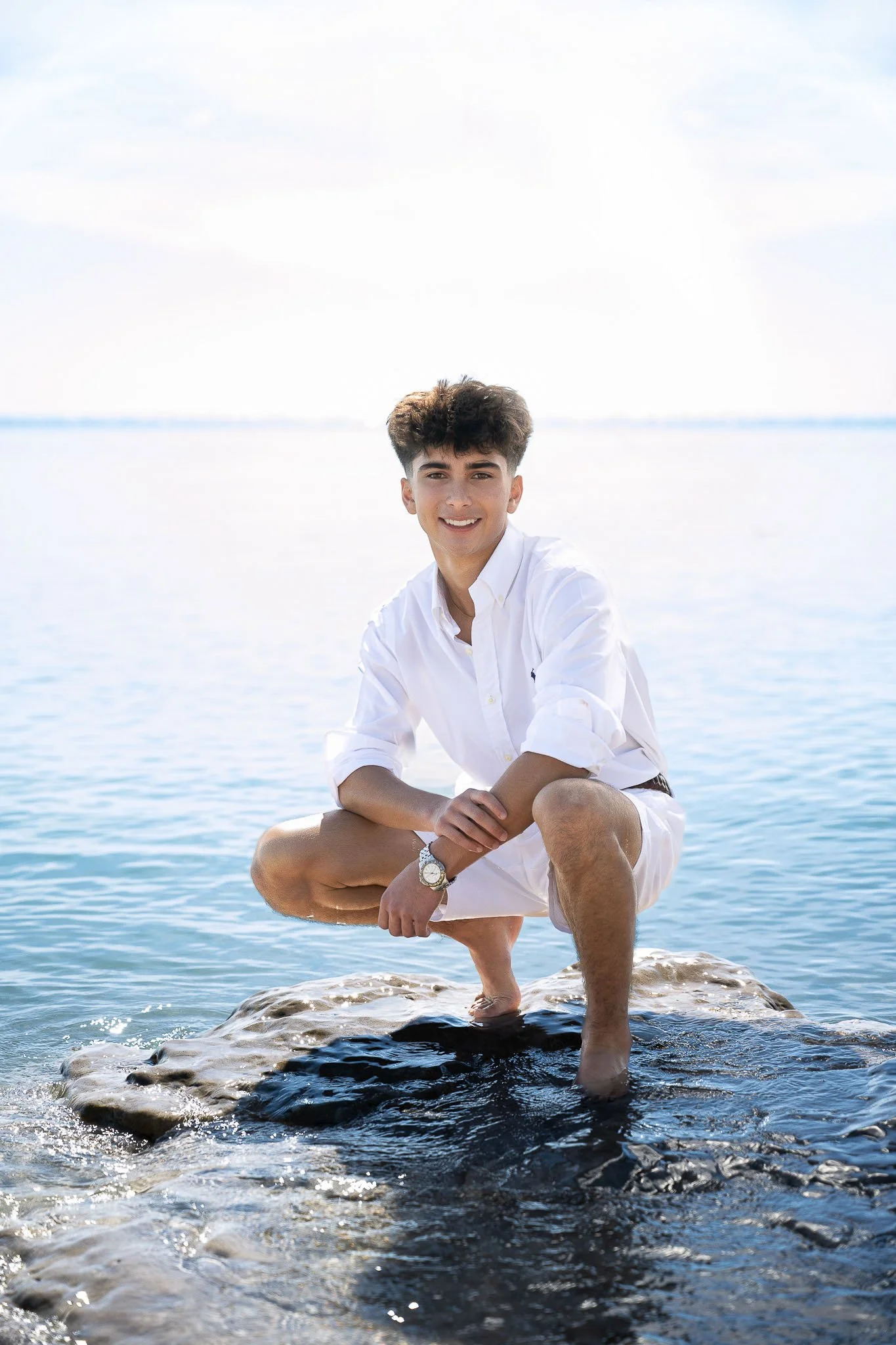 Teen boy crouching barefoot on lakeside rocks, white button-down shirt, smiling at camera with calm lake water and bright open sky behind — Milwaukee senior portrait photography