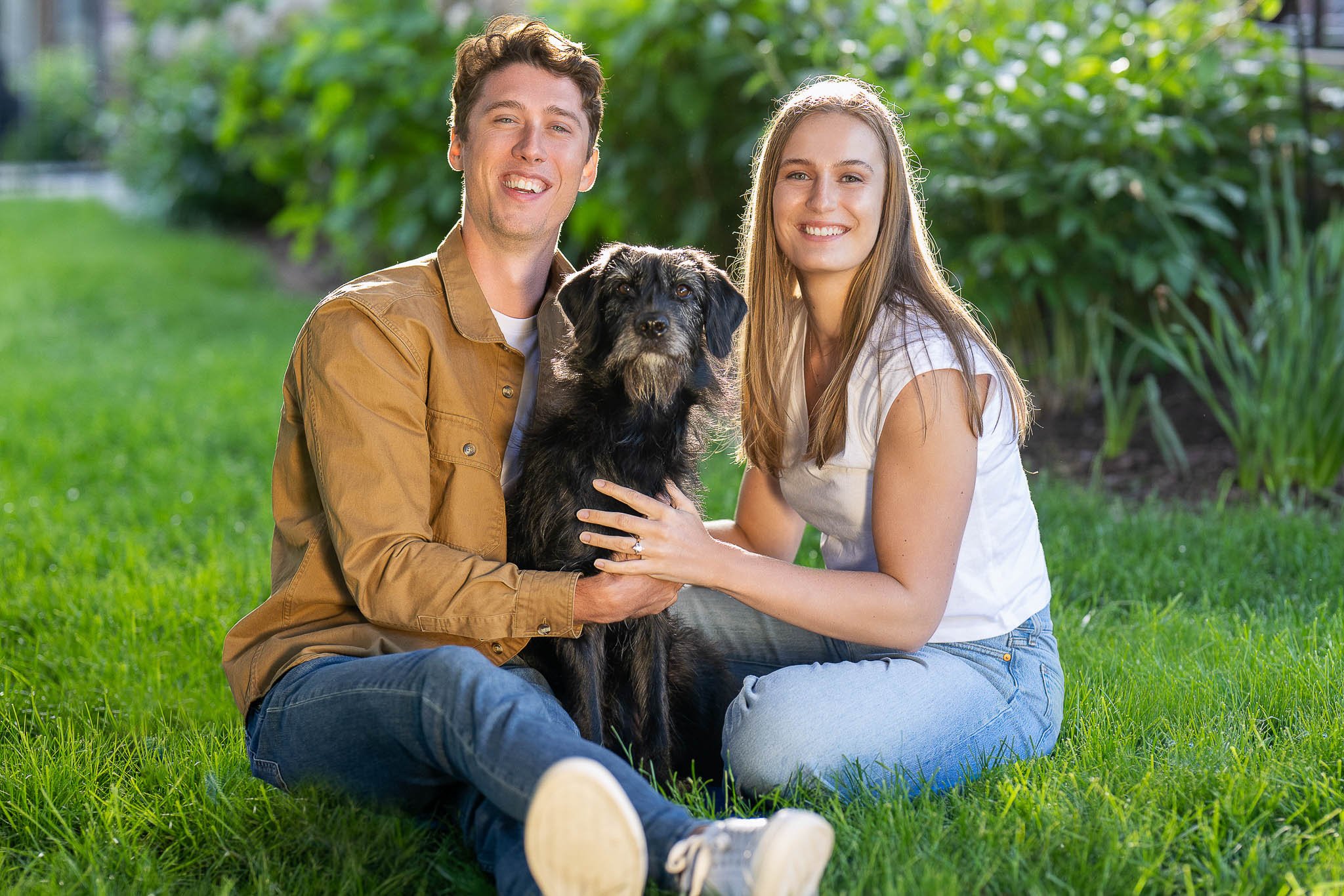 Casual engagement session on lawn, couple sitting together with their black dog, man in tan shirt and jeans, woman in white t-shirt, green garden setting — Milwaukee engagement photography