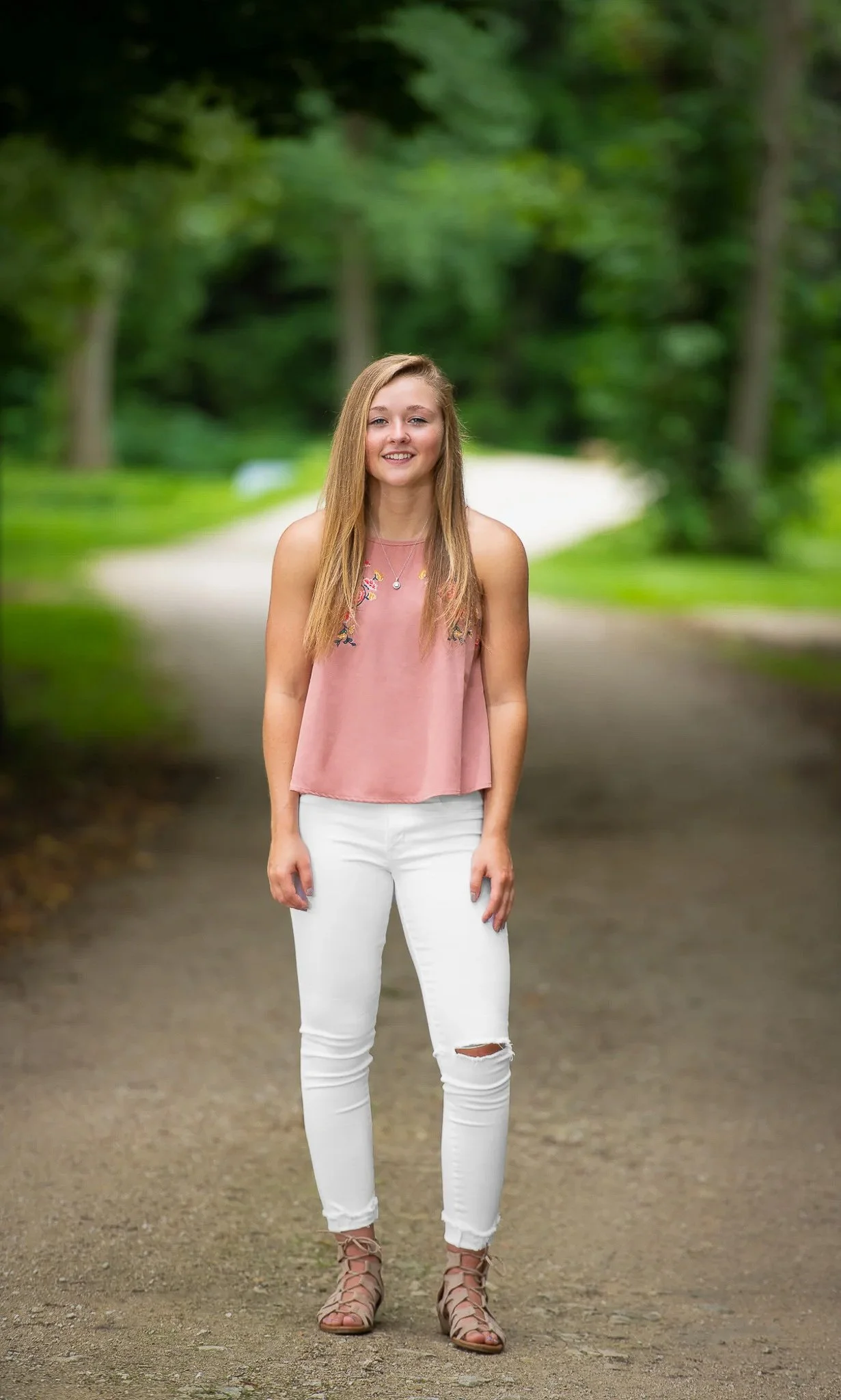 Teen girl standing on park path in pink embroidered sleeveless top, white ripped jeans, and lace-up sandals, lush green trees softly blurred behind — Milwaukee senior portrait photography