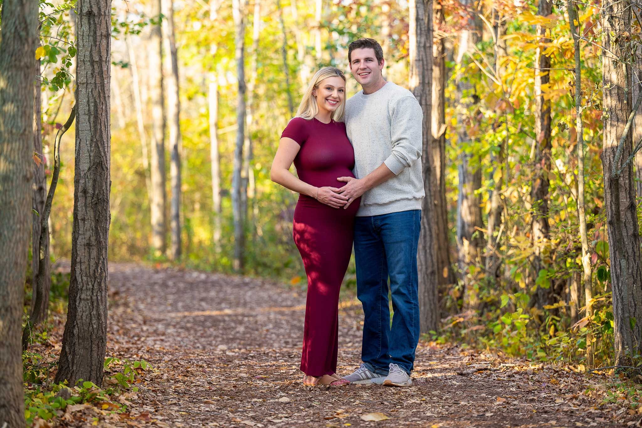 Expecting couple on autumn forest trail, woman in fitted burgundy maxi dress with hand on belly, man in cream sweater and jeans, golden fall foliage surrounding — Milwaukee maternity portrait photography