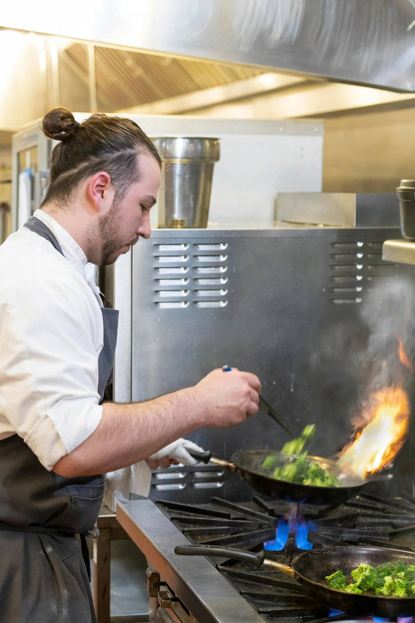 Chef cooking at commercial kitchen station with pan fire and steam rising, professional kitchen equipment in background — Milwaukee culinary event photography