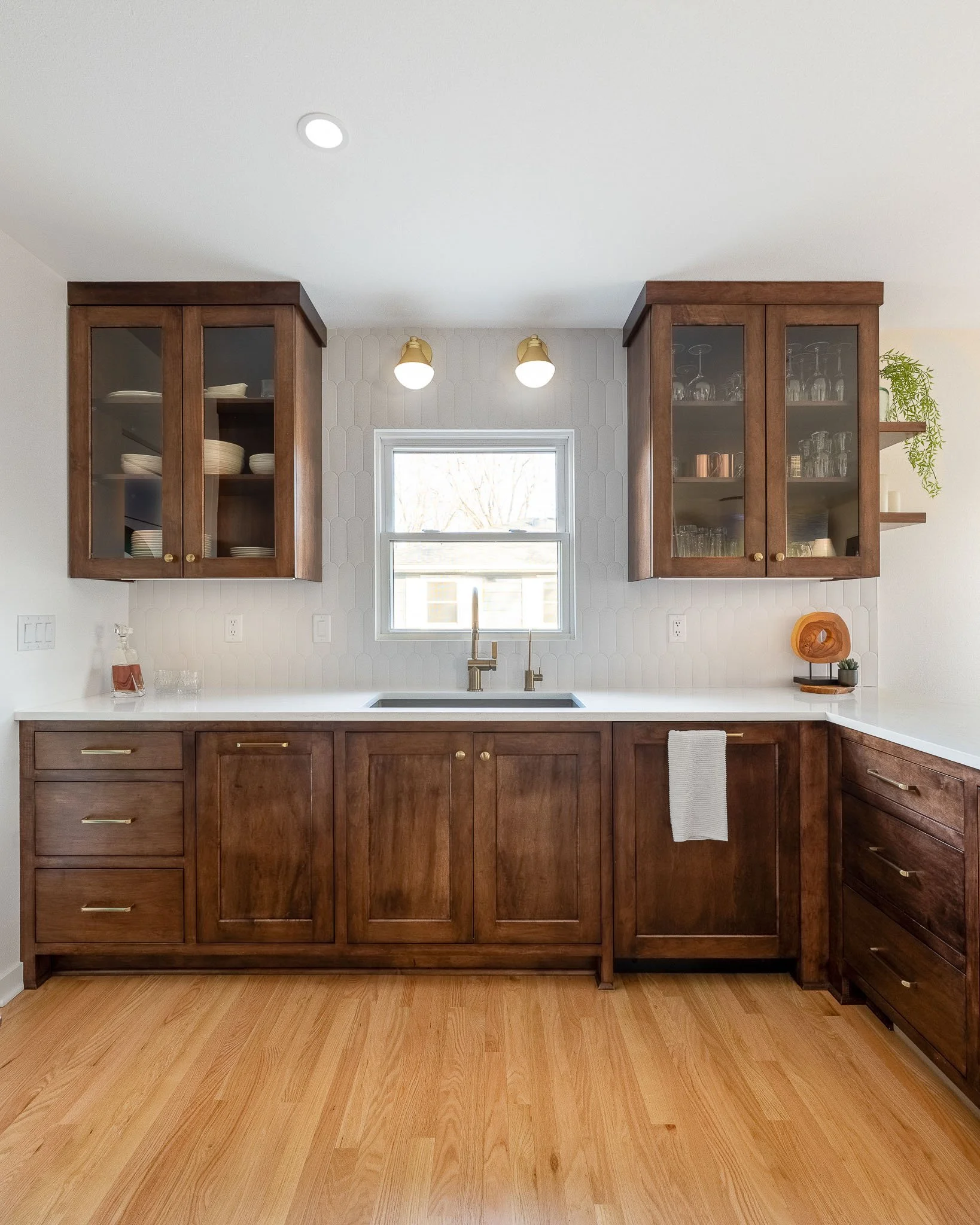 Dark walnut kitchen with glass-front upper cabinets, elongated hexagonal tile backsplash, paired brass sconces, and white quartz countertops — Milwaukee Macpherson residential interior photography