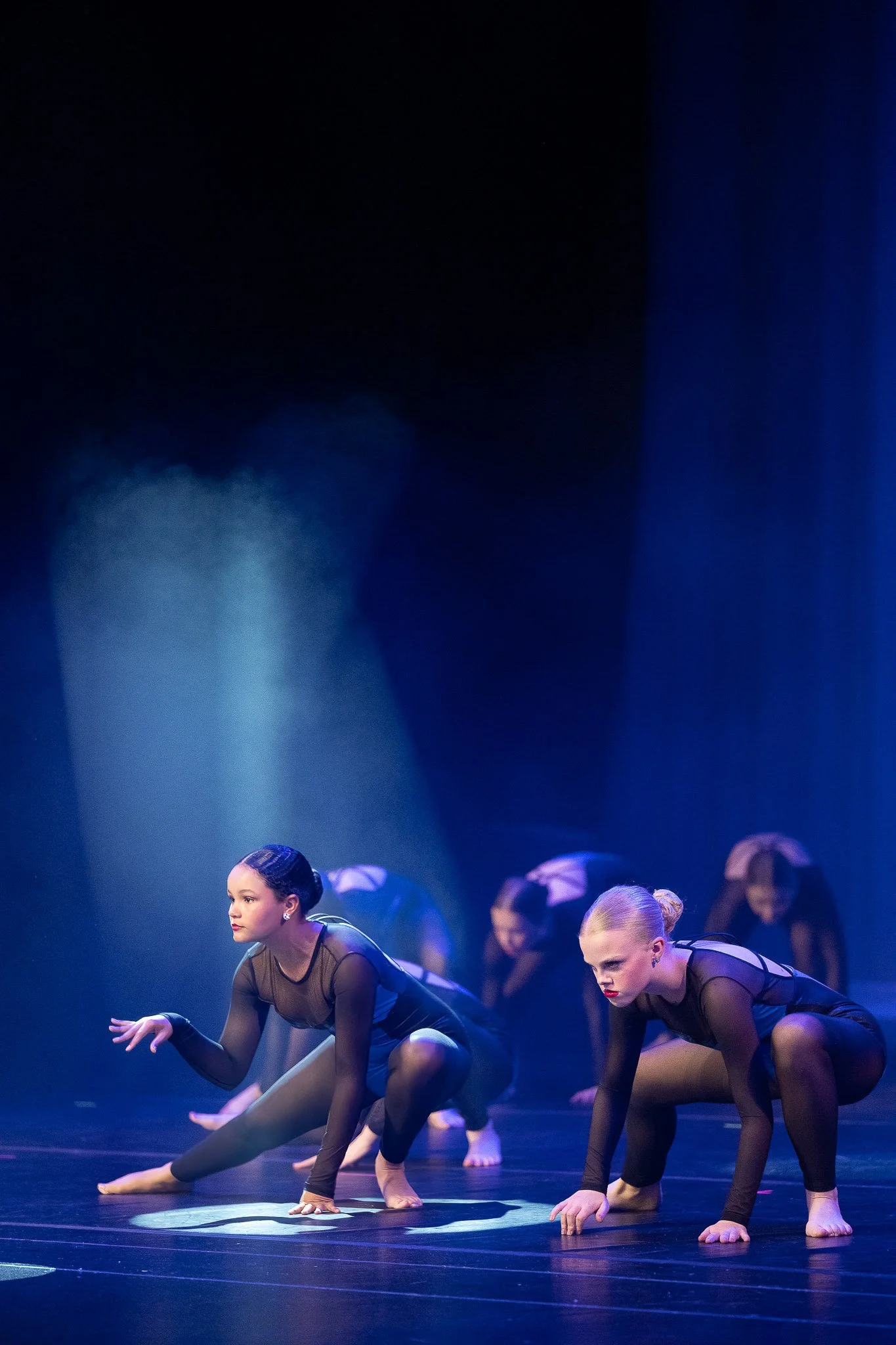 Contemporary dance performance, two young dancers in black mesh costumes crouching low on stage with dramatic blue spotlight and stage fog — Milwaukee event photography