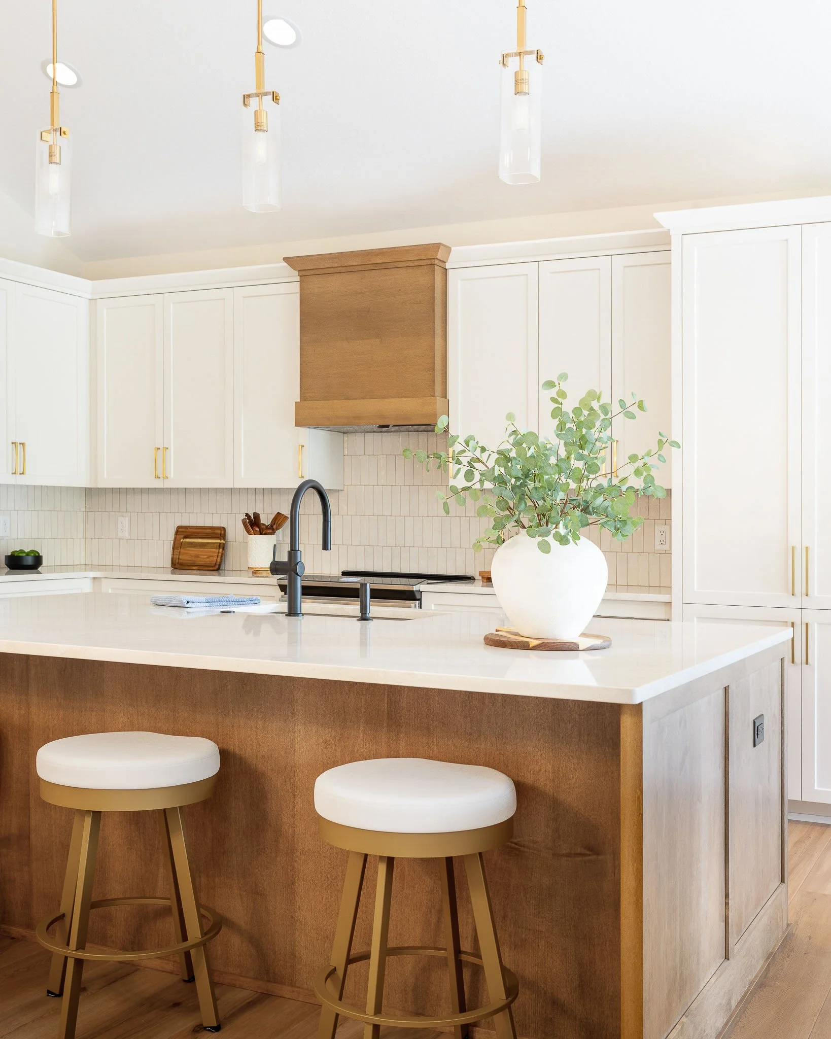 White transitional kitchen with warm wood island, wood range hood, brass hardware, gold cylinder pendants, and eucalyptus arrangement — Milwaukee residential interior photography