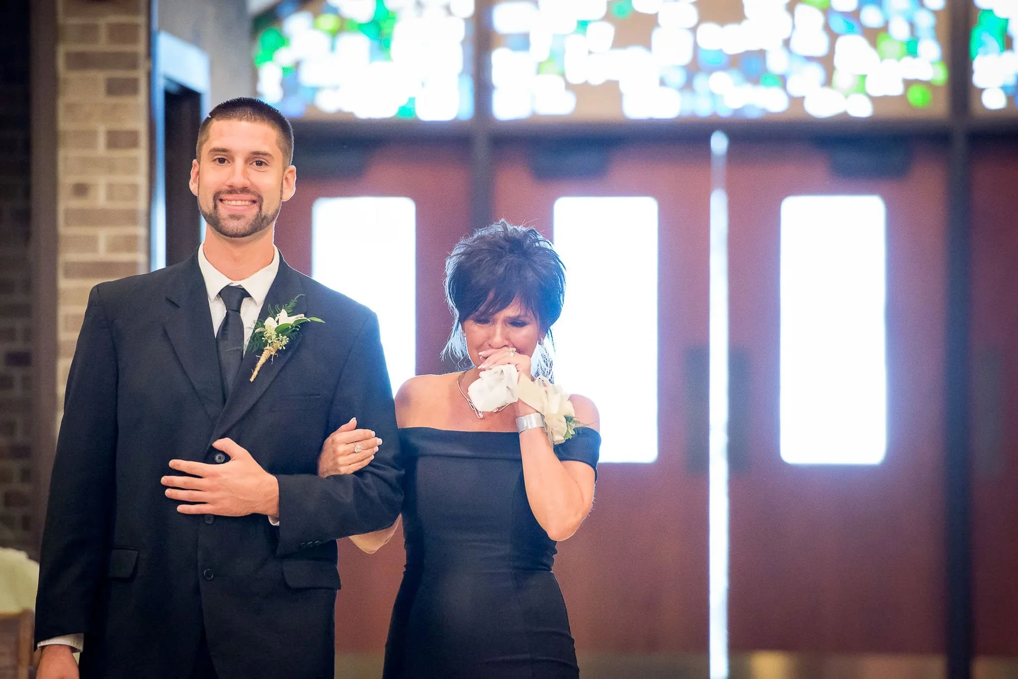 Groom walking emotional mother down church aisle with stained glass windows and wooden doors glowing behind them — Milwaukee wedding photography