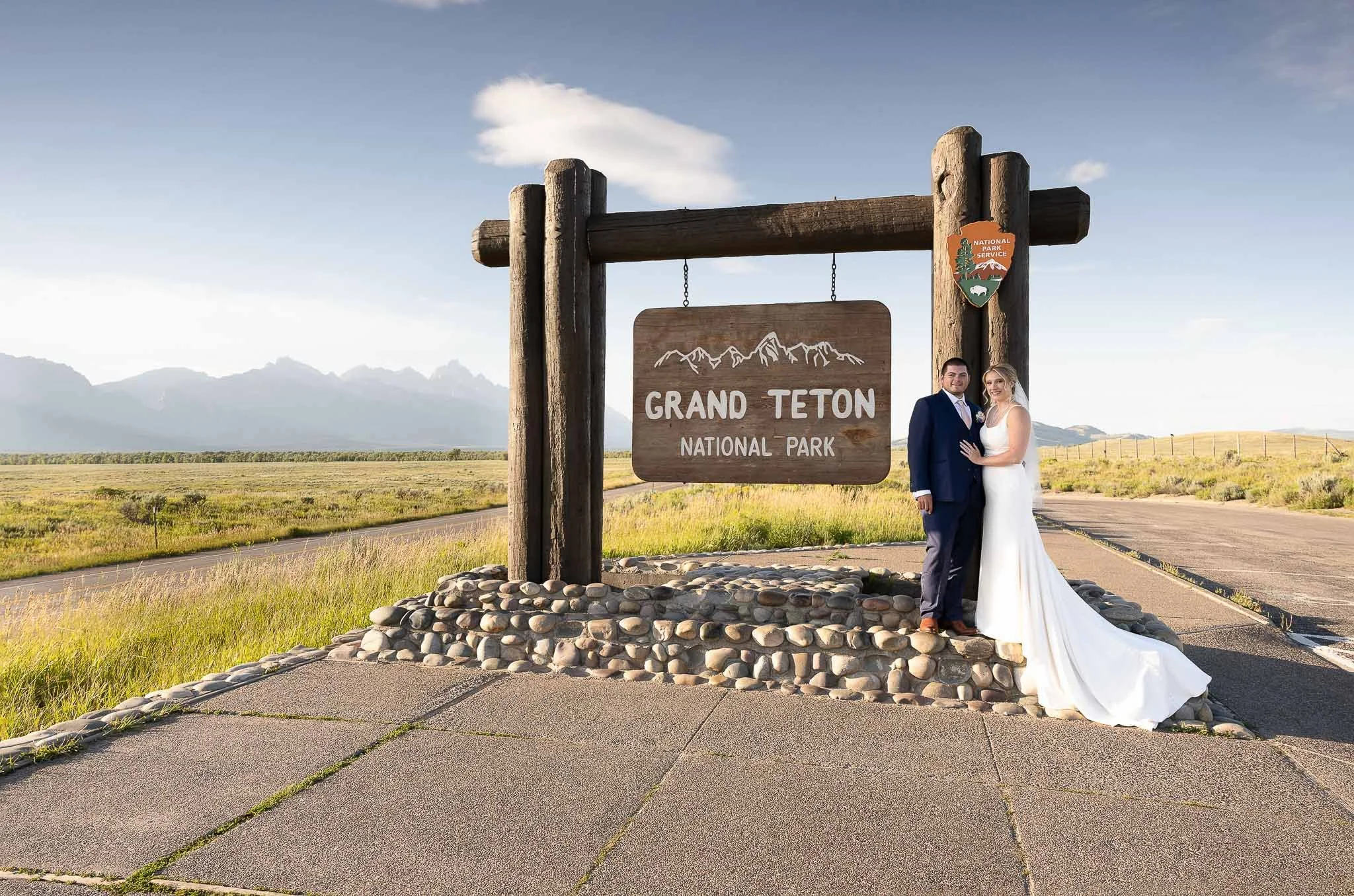 Couple portrait at Grand Teton National Park entrance sign, groom in navy suit, bride in simple white gown, mountain peaks and blue sky — Jackson Hole Wyoming wedding photography