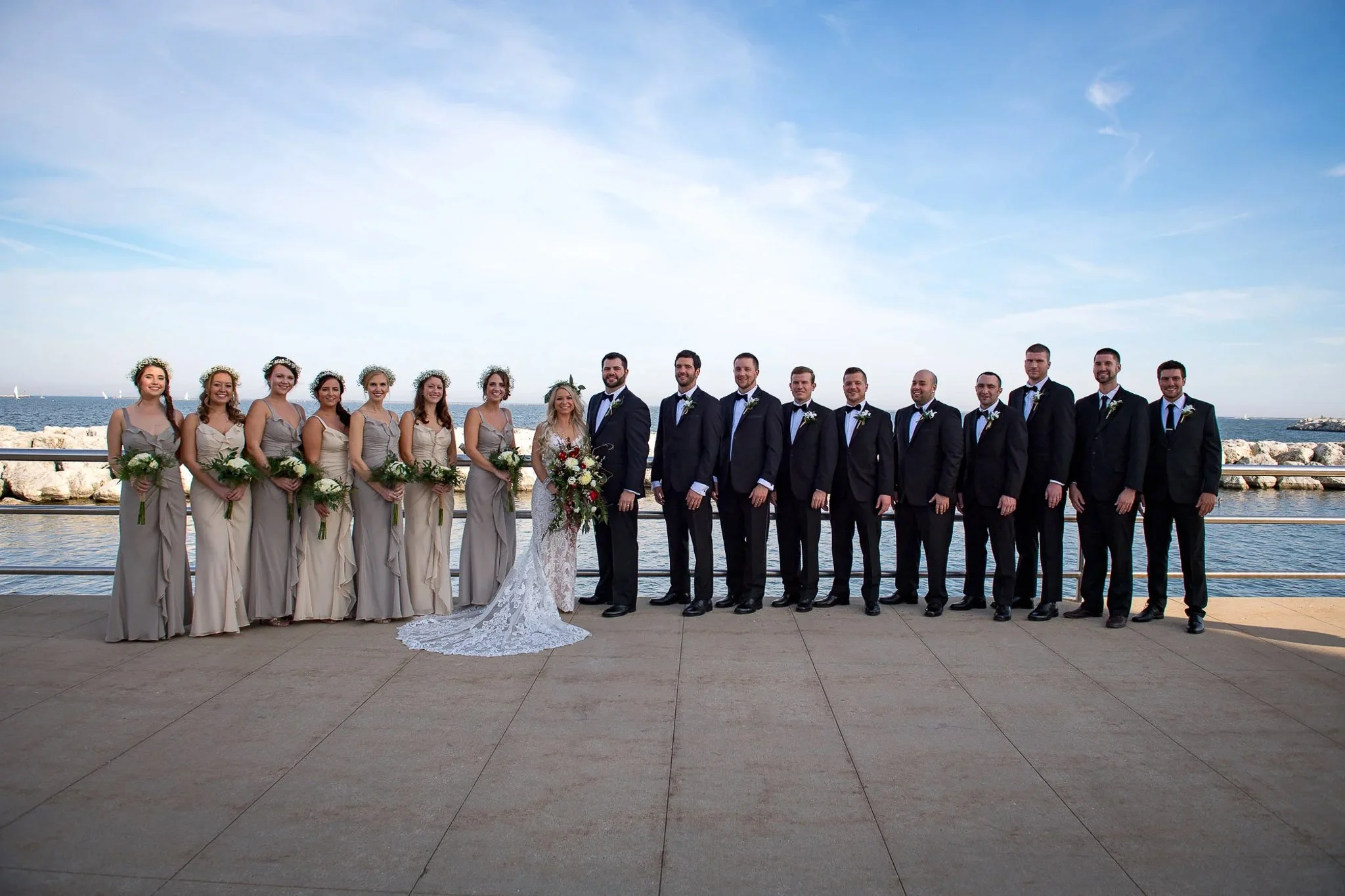 Full wedding party on Milwaukee lakefront promenade, champagne-taupe bridesmaids with flower crowns, groomsmen in black tuxedos, Lake Michigan behind — Milwaukee wedding photography
