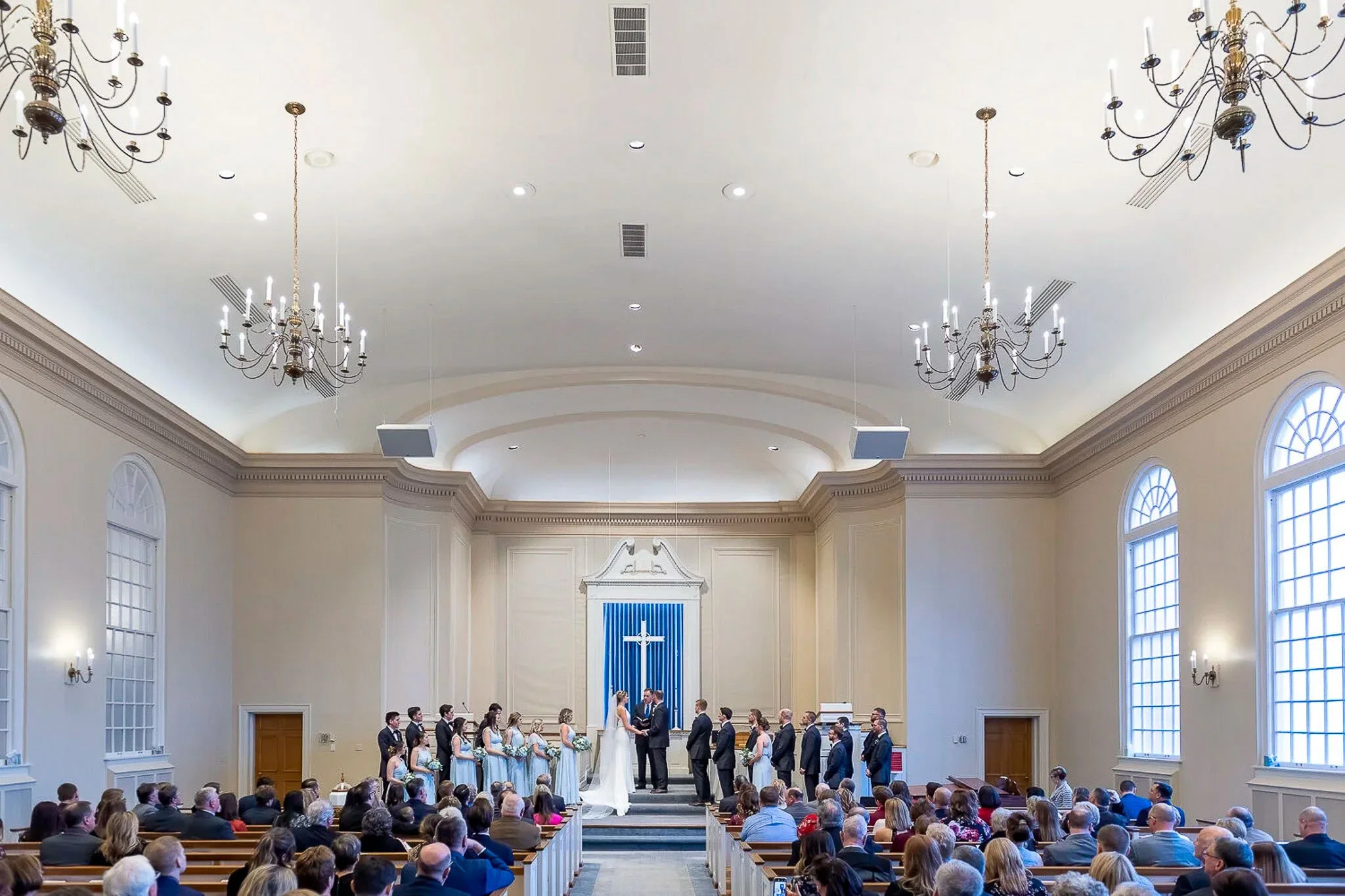 Wide interior view of white church ceremony with candle chandeliers, blue fabric cross backdrop, wedding party flanking couple at the altar with full congregation — Milwaukee wedding photography