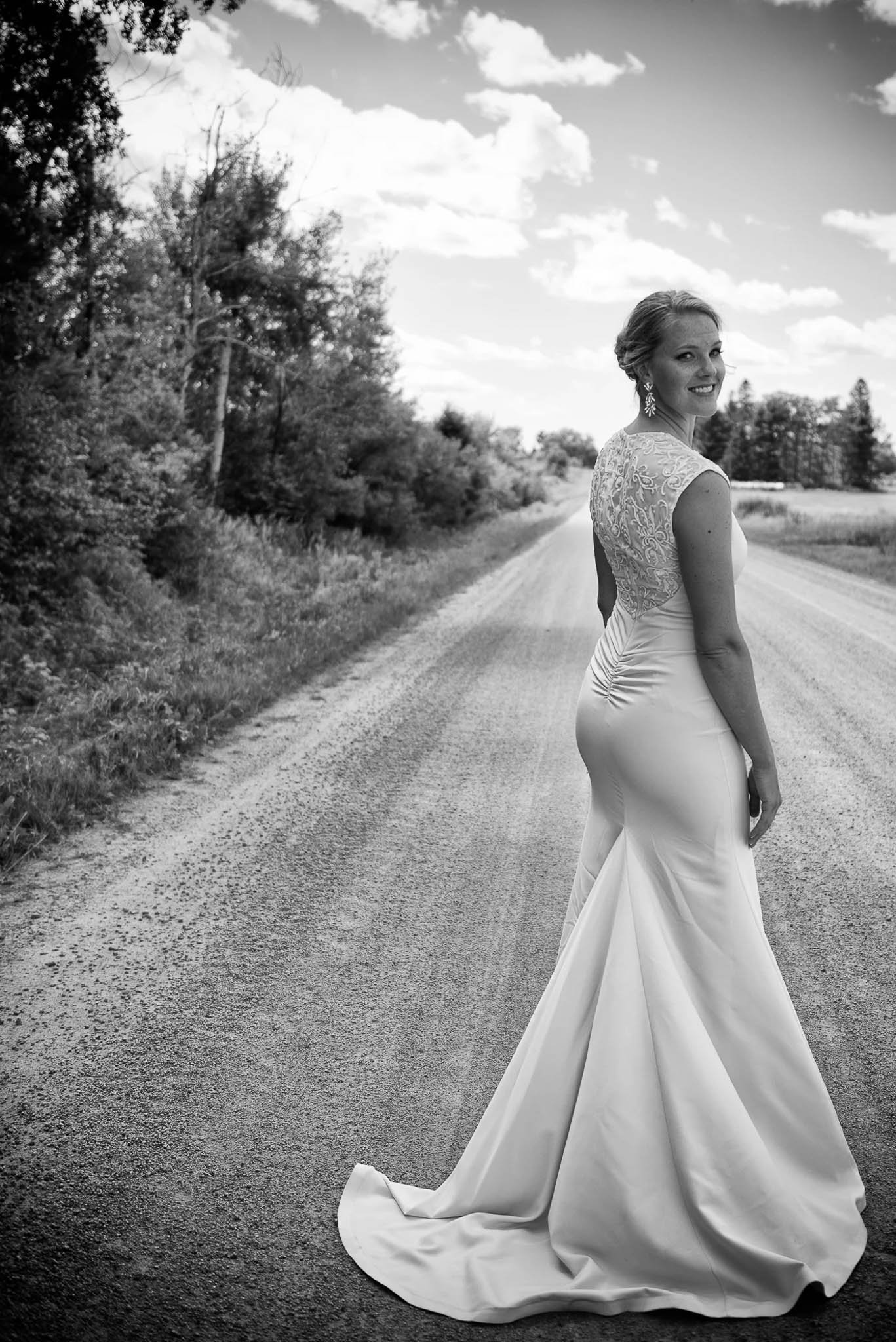 Black and white bride portrait on rural country road looking back over shoulder, fitted cap-sleeve lace gown with long train, tree-lined road vanishing into distance — Milwaukee wedding photography