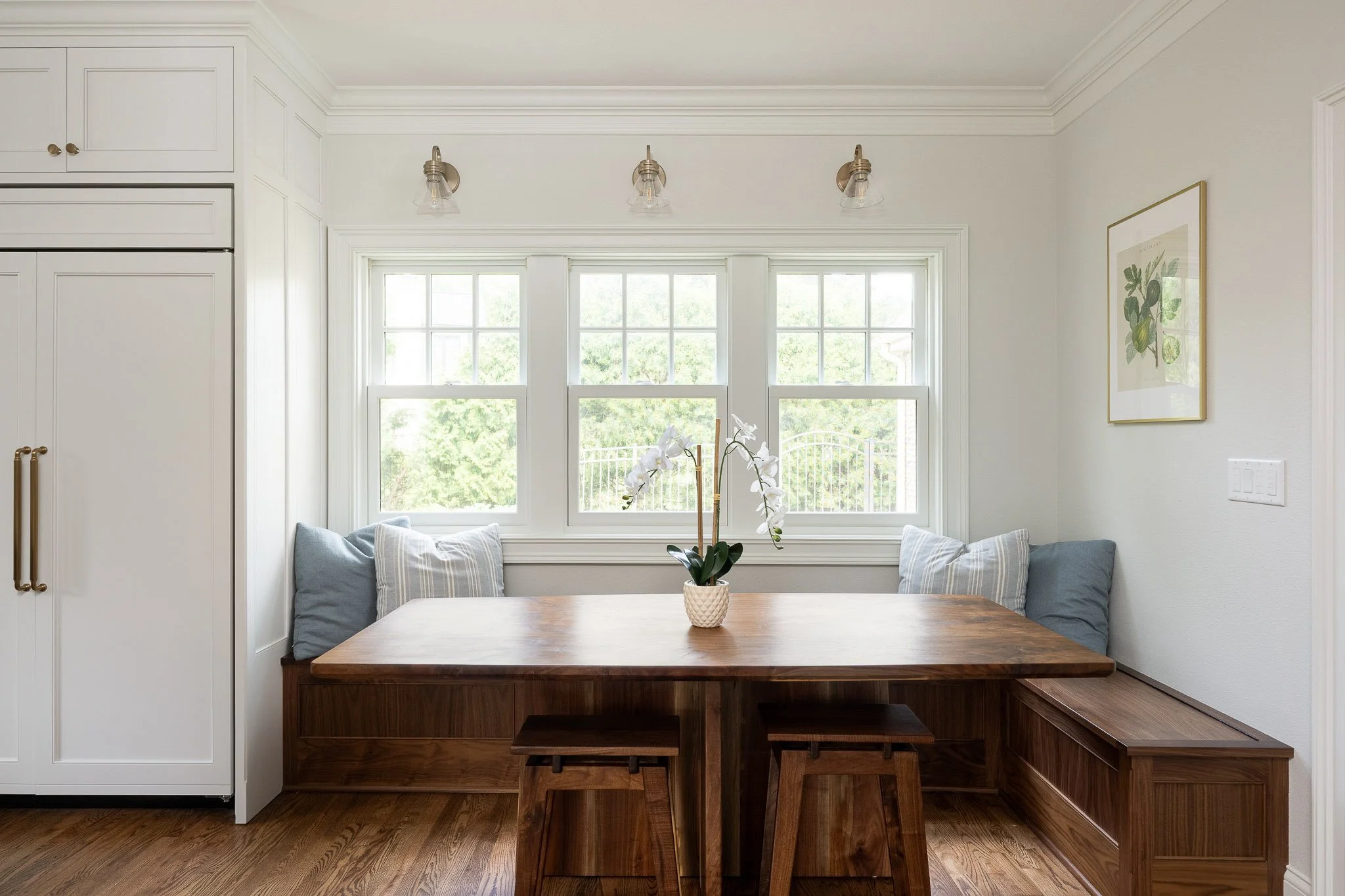 Walnut L-shaped breakfast nook with built-in bench seating, walnut dining table, orchid centerpiece, brass sconces, and garden window views — Milwaukee residential interior photography