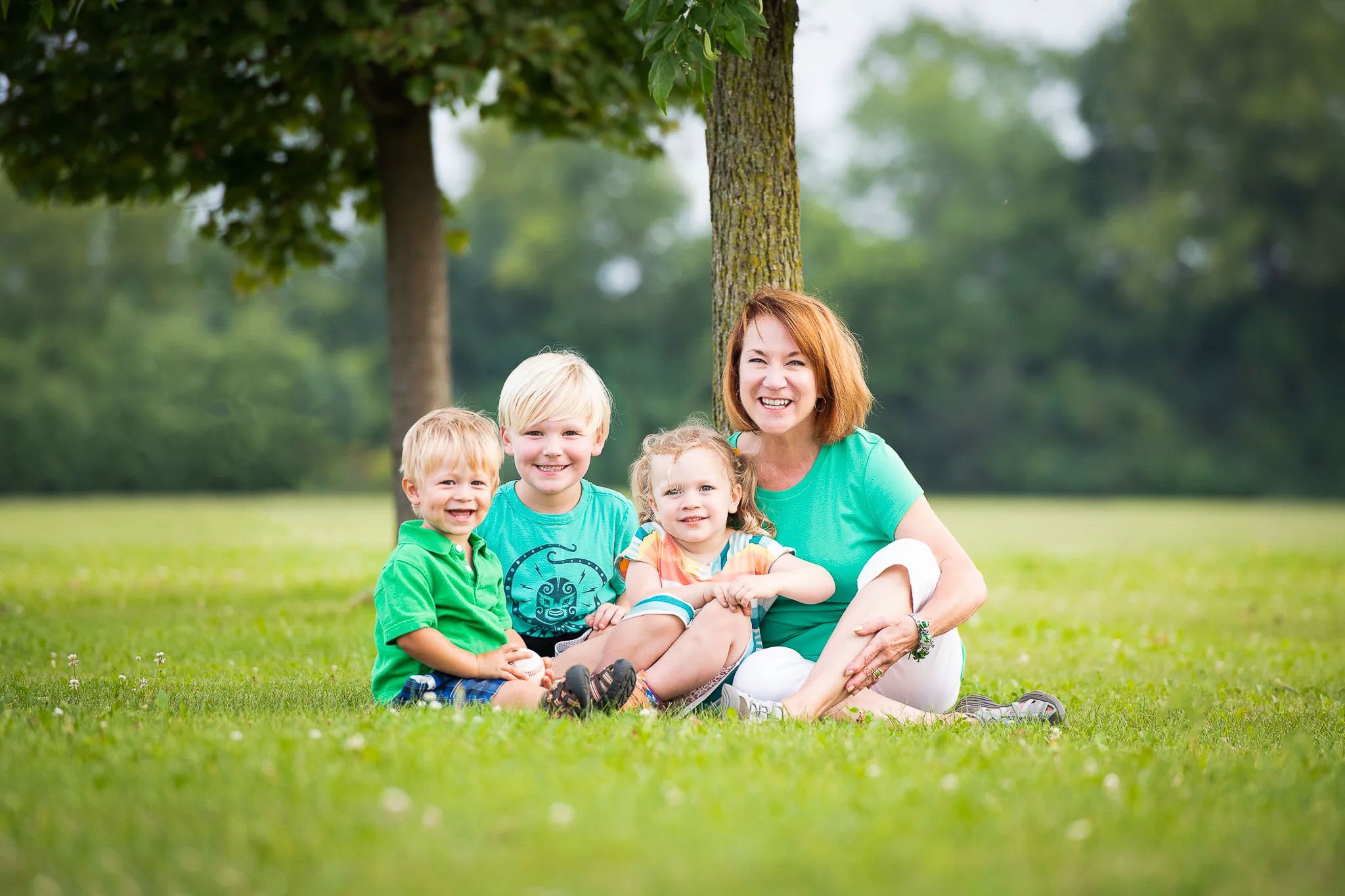 Grandmother with three young grandchildren sitting together on green lawn under a large tree, all in coordinated green outfits, smiling at camera — Milwaukee family portrait photography