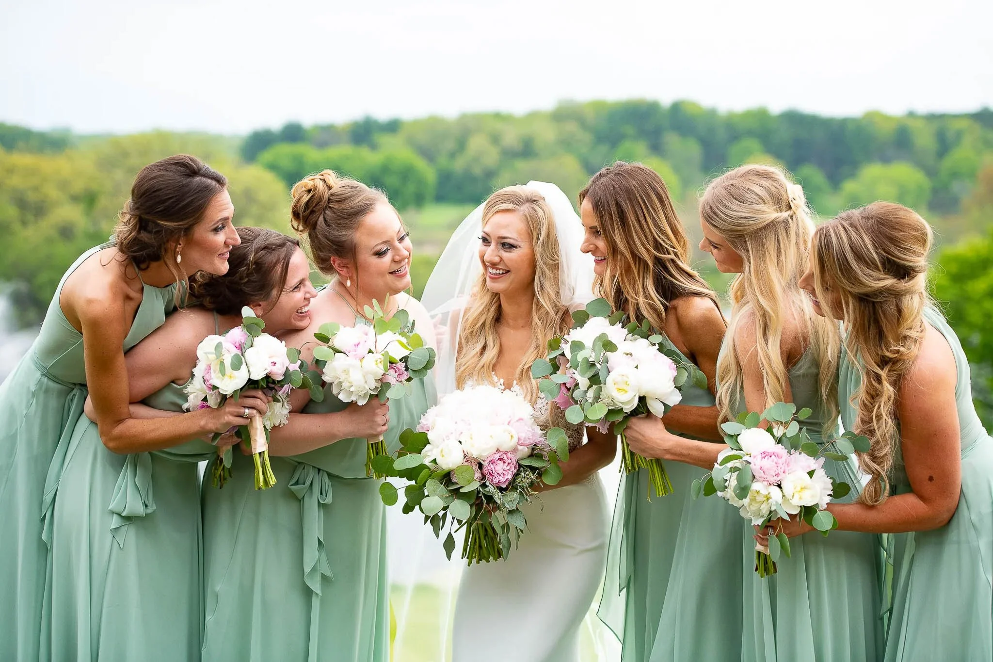 Mint green bridesmaids surrounding laughing bride in a huddle, white and pink peony bouquets with eucalyptus, green rolling hills in background — Milwaukee wedding photography