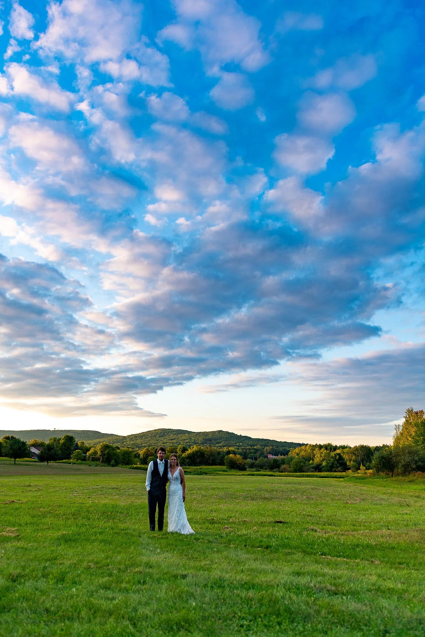 Couple standing together in vast green meadow under dramatic cloudscape, groom in navy vest, bride in fitted lace gown, rolling wooded hills on the horizon — Milwaukee wedding photography