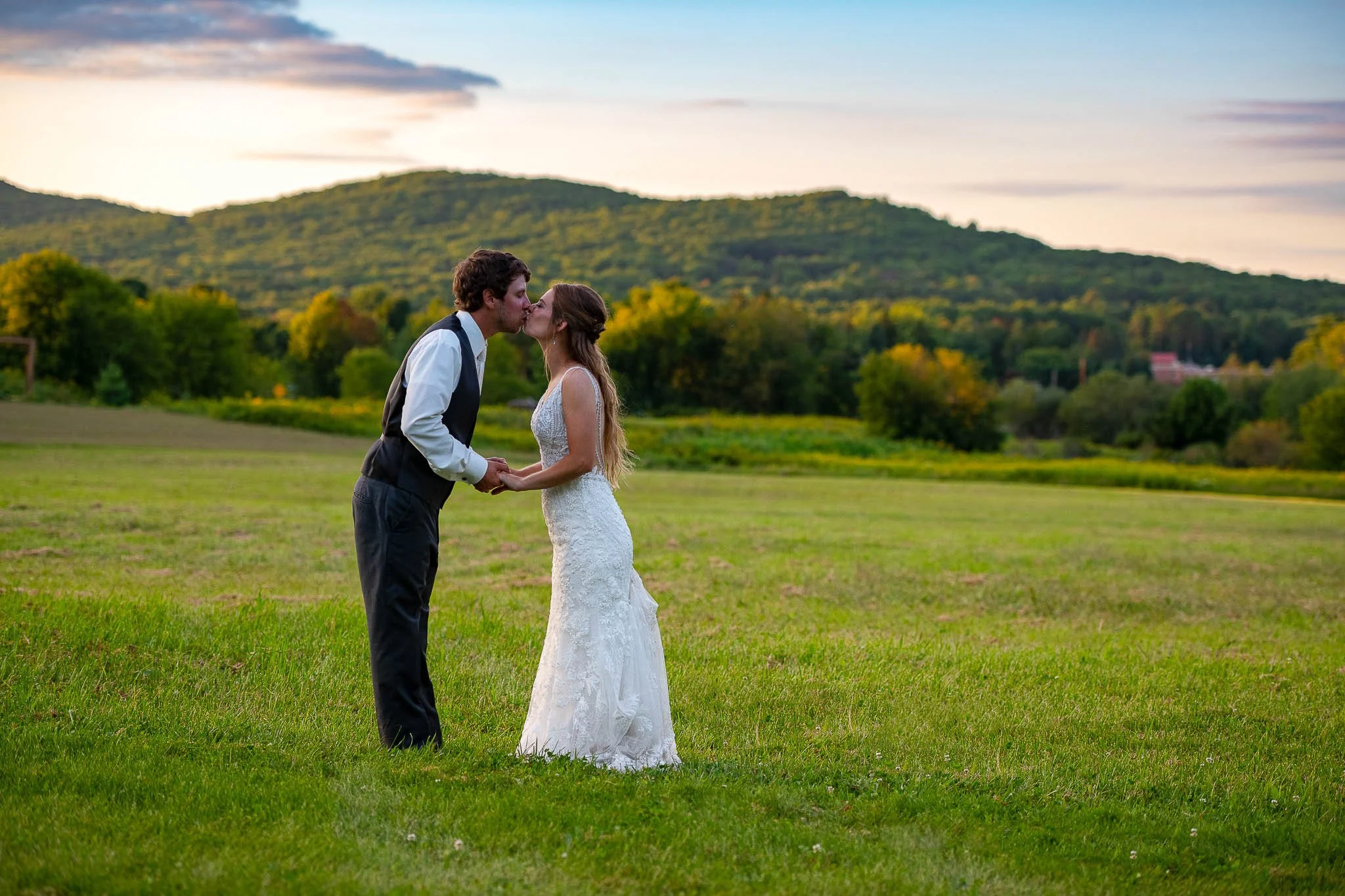 Couple kissing in expansive green meadow at sunset, groom in dark charcoal vest, bride in fitted lace gown, rolling wooded hills under glowing sky — Milwaukee wedding photography