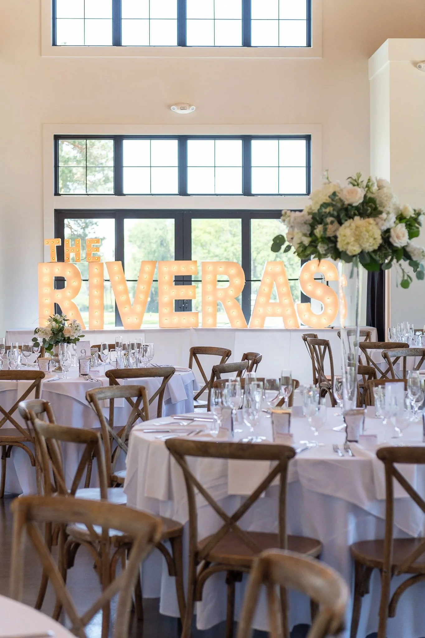Bright reception hall with illuminated "THE RIVERAS" marquee letter sign, wood cross-back chairs, white tablecloths, and tall white floral centerpiece with hydrangeas — Milwaukee wedding photography