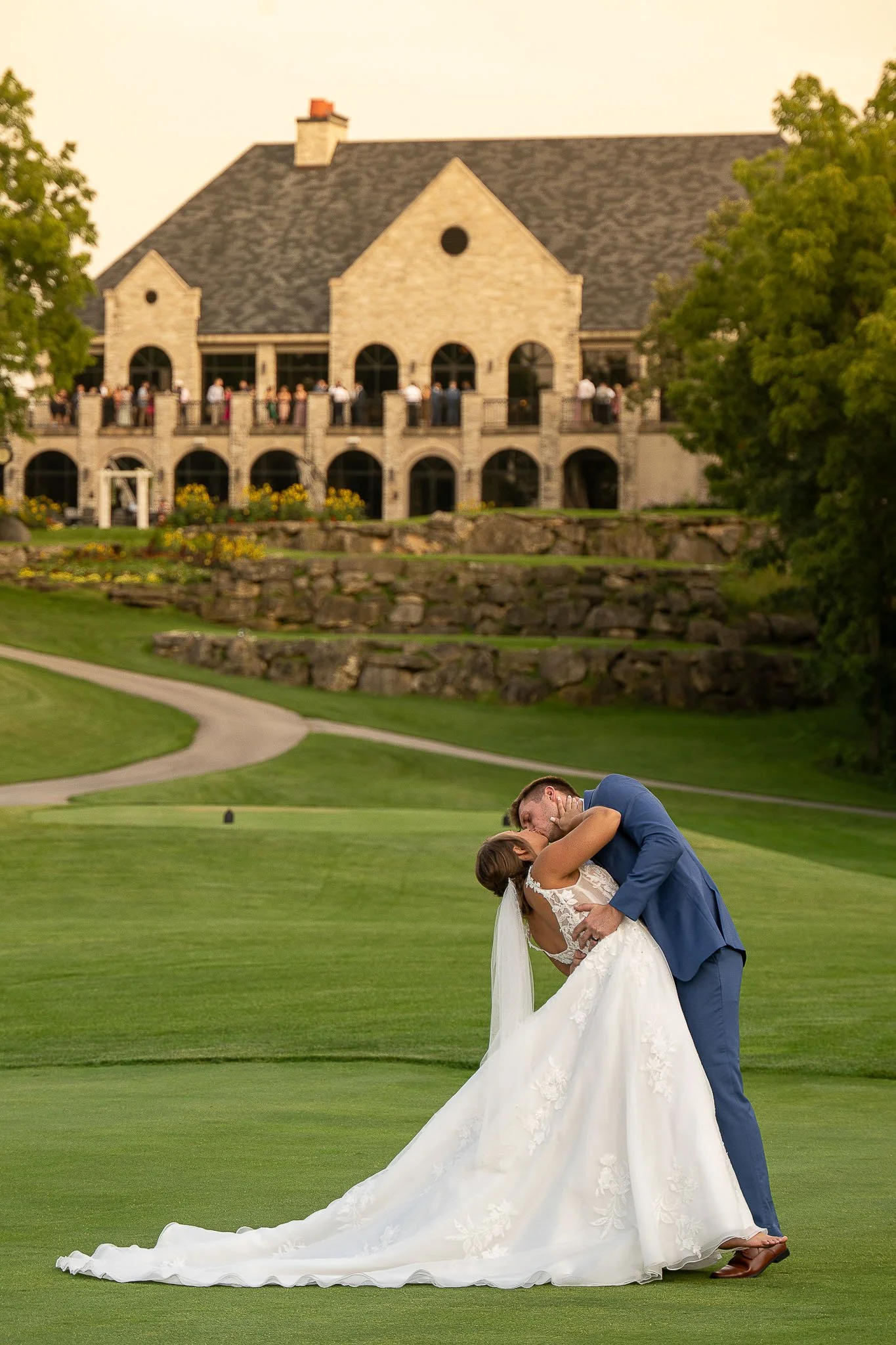 Golf green dip-kiss at golden hour, groom in blue suit, bride in lace ball gown with long cathedral train, stone chateau clubhouse with guests watching from balcony — Milwaukee wedding photography