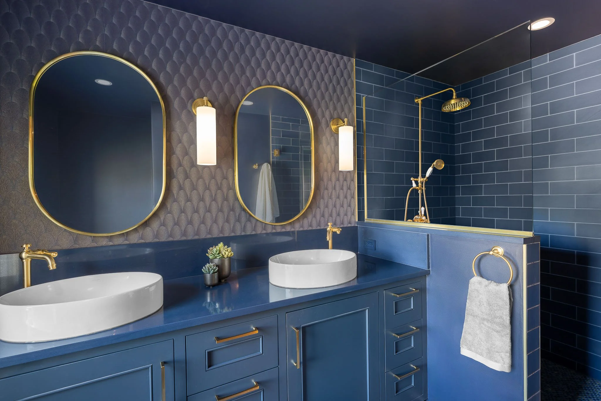 Bold cobalt blue bathroom with dual vessel sinks, blue vanity, gold oval mirrors, scallop patterned wallpaper, and blue subway tile walk-in shower — Milwaukee residential interior photography