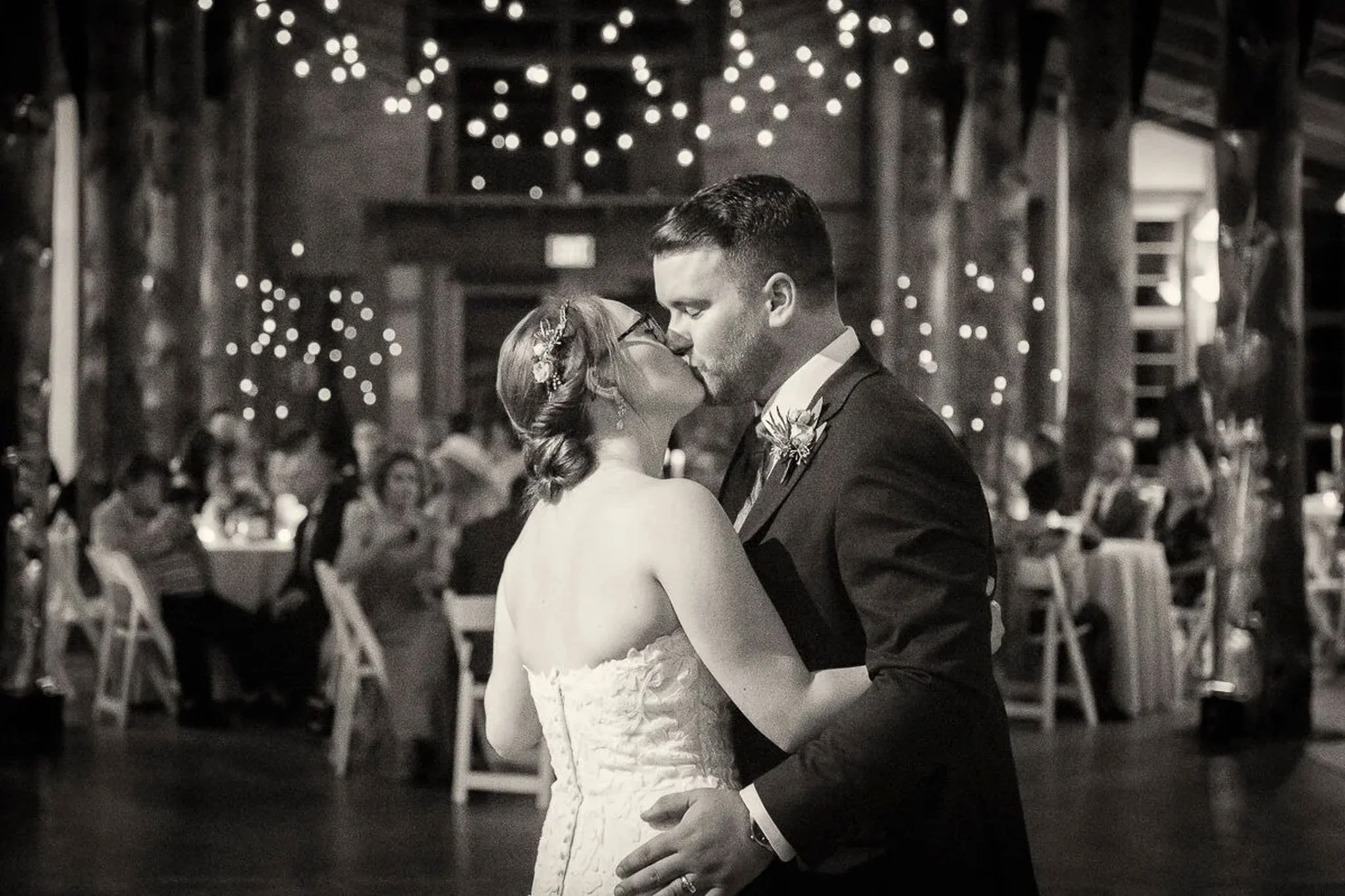 Black and white first dance in rustic barn reception hall with bokeh string lights overhead, guests seated at candlelit tables in background — Milwaukee wedding photography