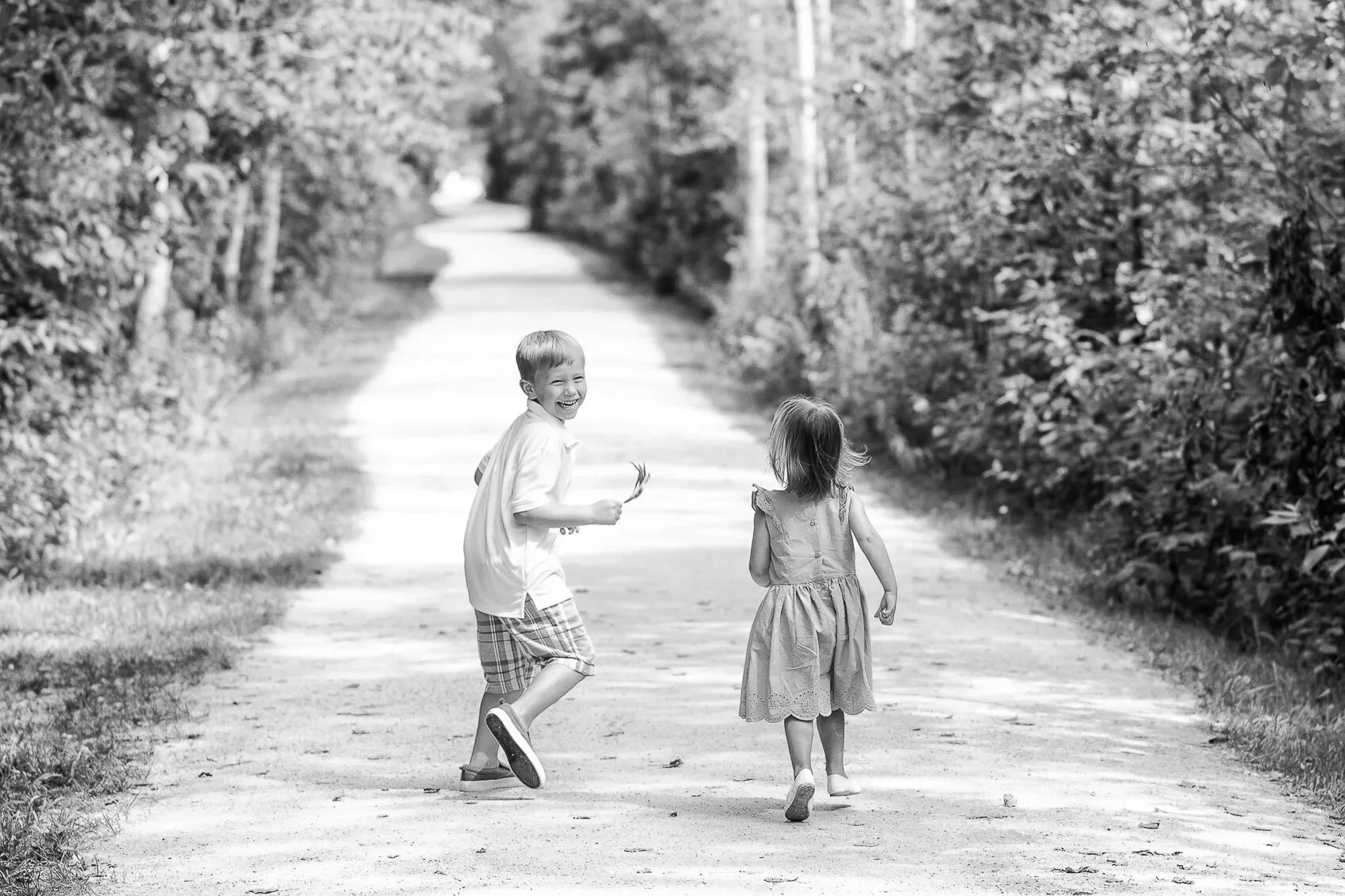 Black and white portrait of two young siblings playing on a sunlit tree-lined dirt road, laughing boy in plaid shorts turning back, little girl in ruffled dress walking ahead — Milwaukee children's portrait photography
