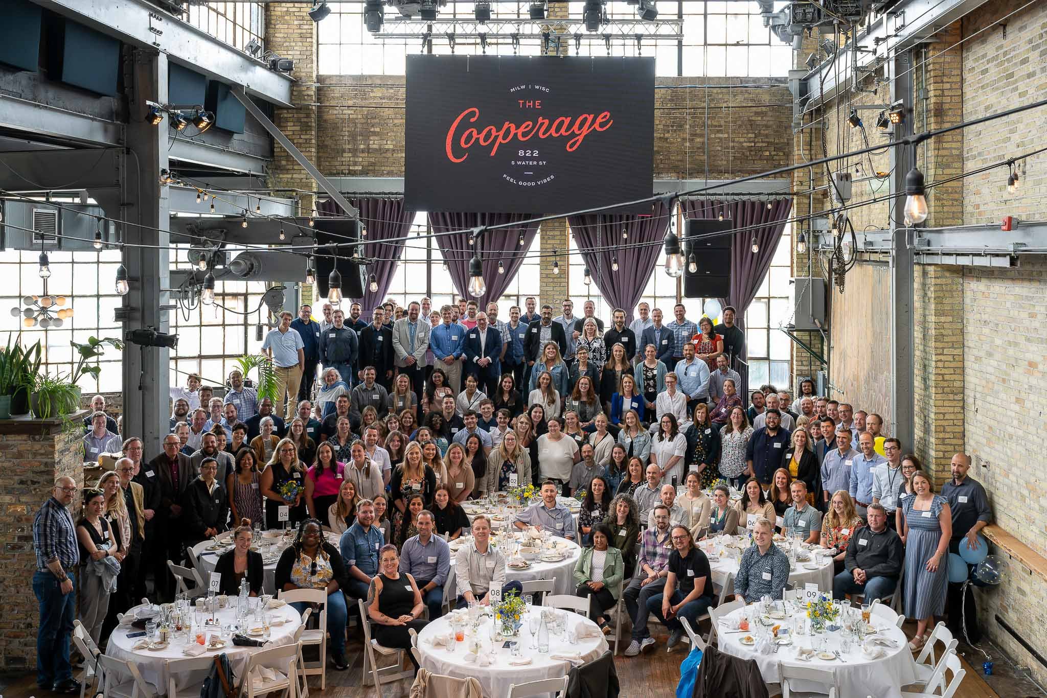 Large corporate group photo at The Cooperage Milwaukee, brick industrial interior with glowing "The Cooperage" neon sign overhead, white round tables in foreground — Milwaukee corporate event photography