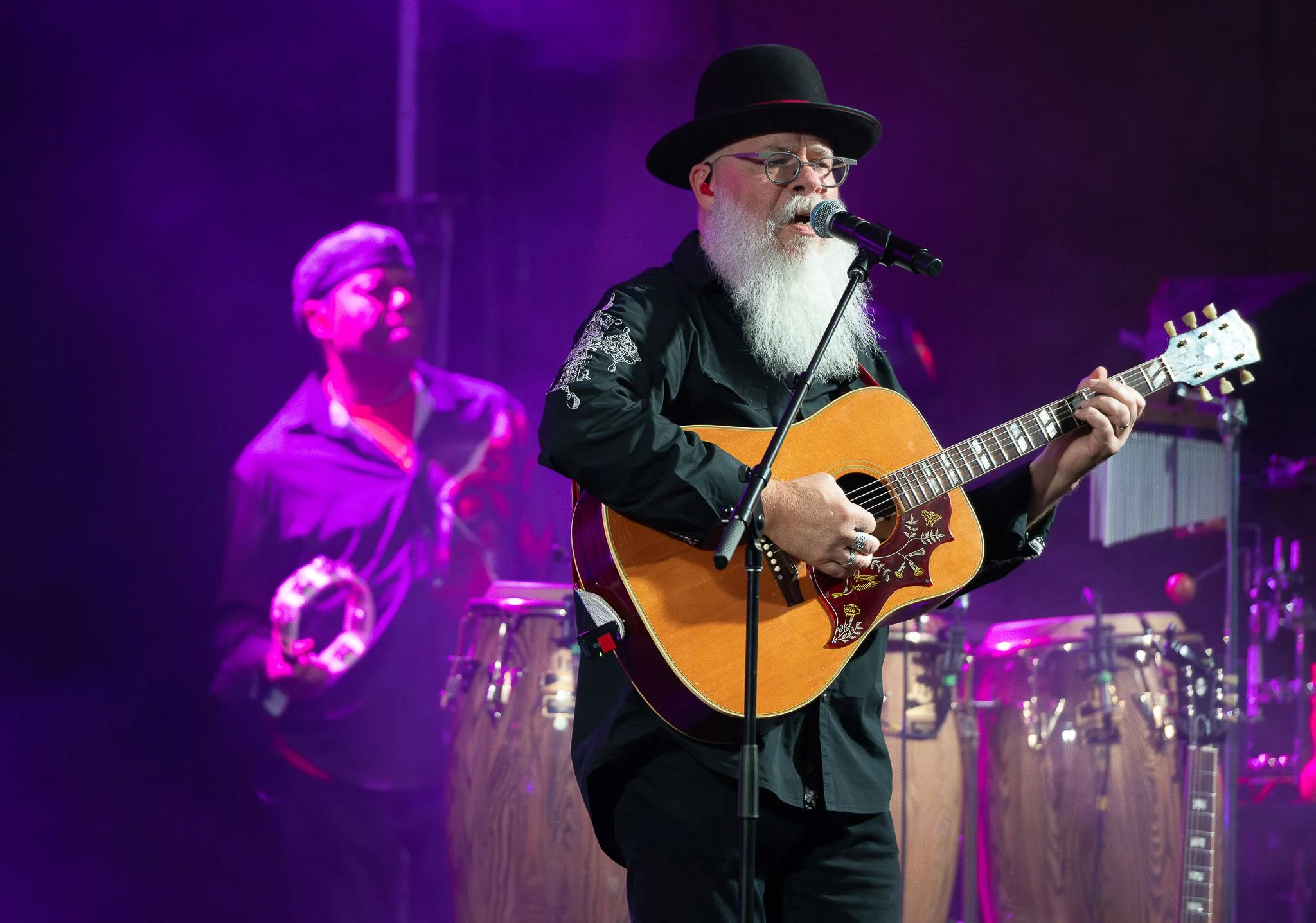 Close-up of male guitarist with long white beard and black bowler hat playing acoustic guitar on stage, purple stage lighting — Milwaukee event band photography