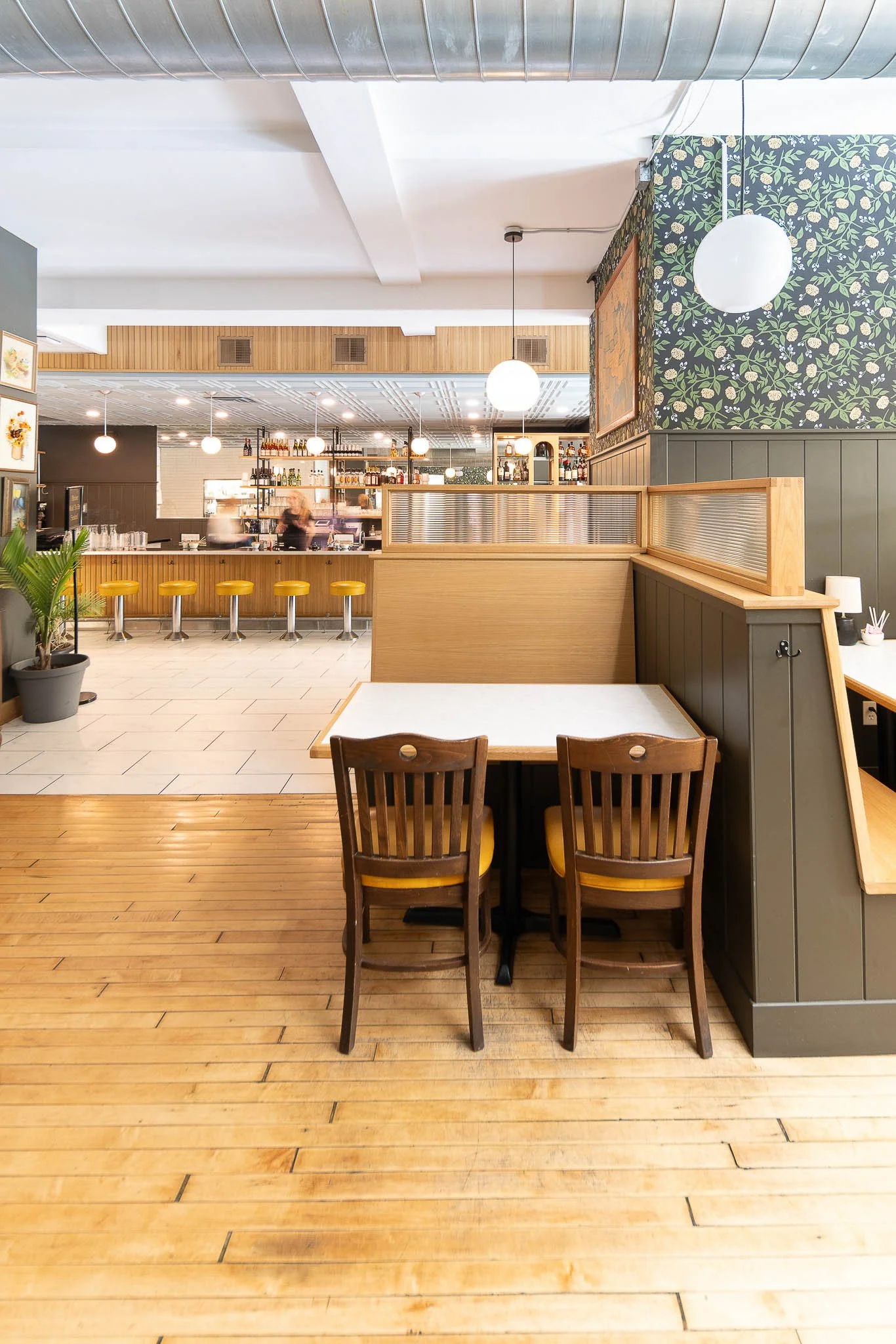 Interior of a restaurant with a small table and chairs in the foreground, a bar with yellow stools in the middle, and a counter with liquor bottles in the background. The decor includes a floral wallpaper, wooden paneling, pendant globe lights, and a potted plant.