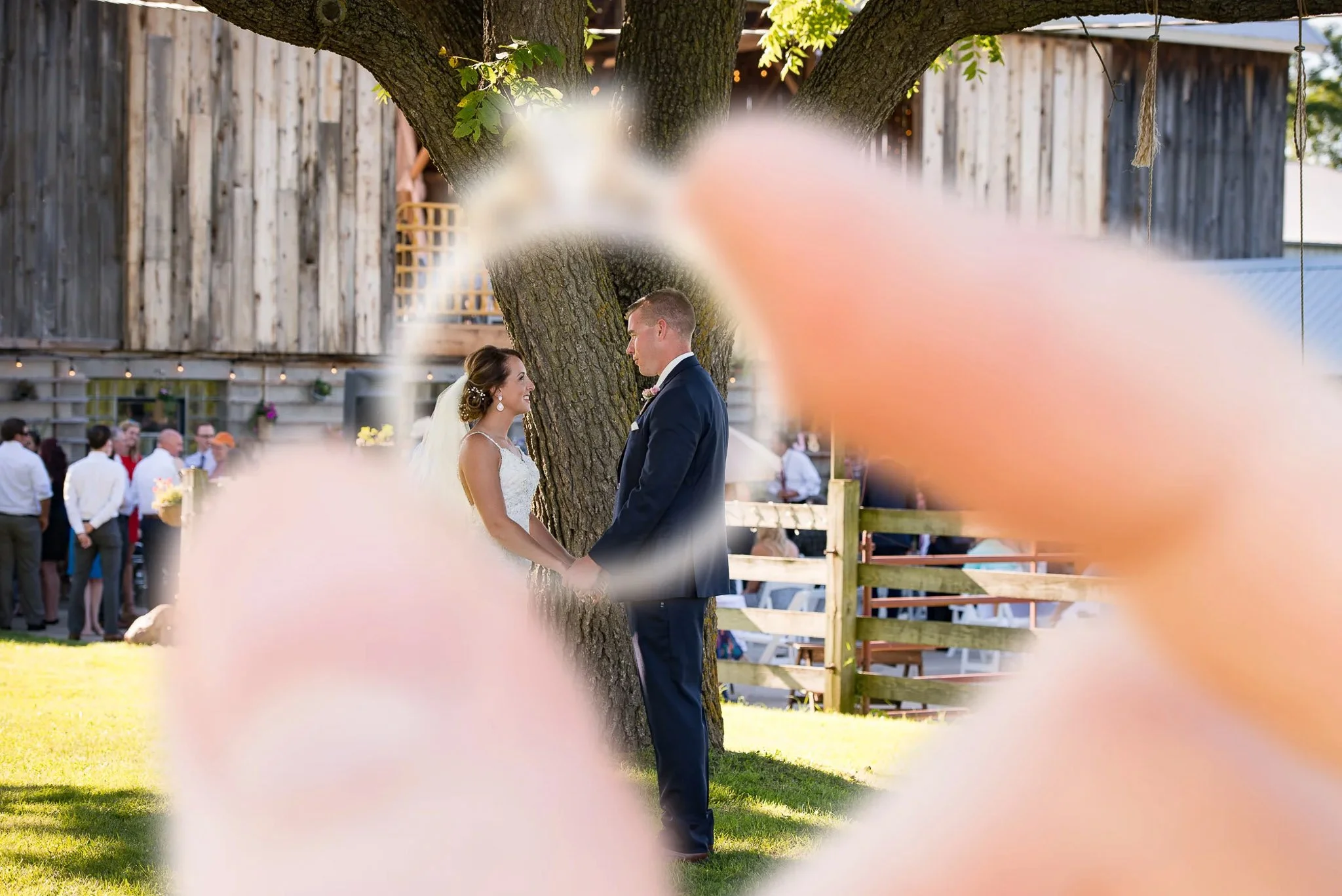 Outdoor barn ceremony framed through bokeh pink flowers in foreground, couple holding hands under large oak tree, navy suit groom, white lace bride, string lights in background — Milwaukee wedding photography